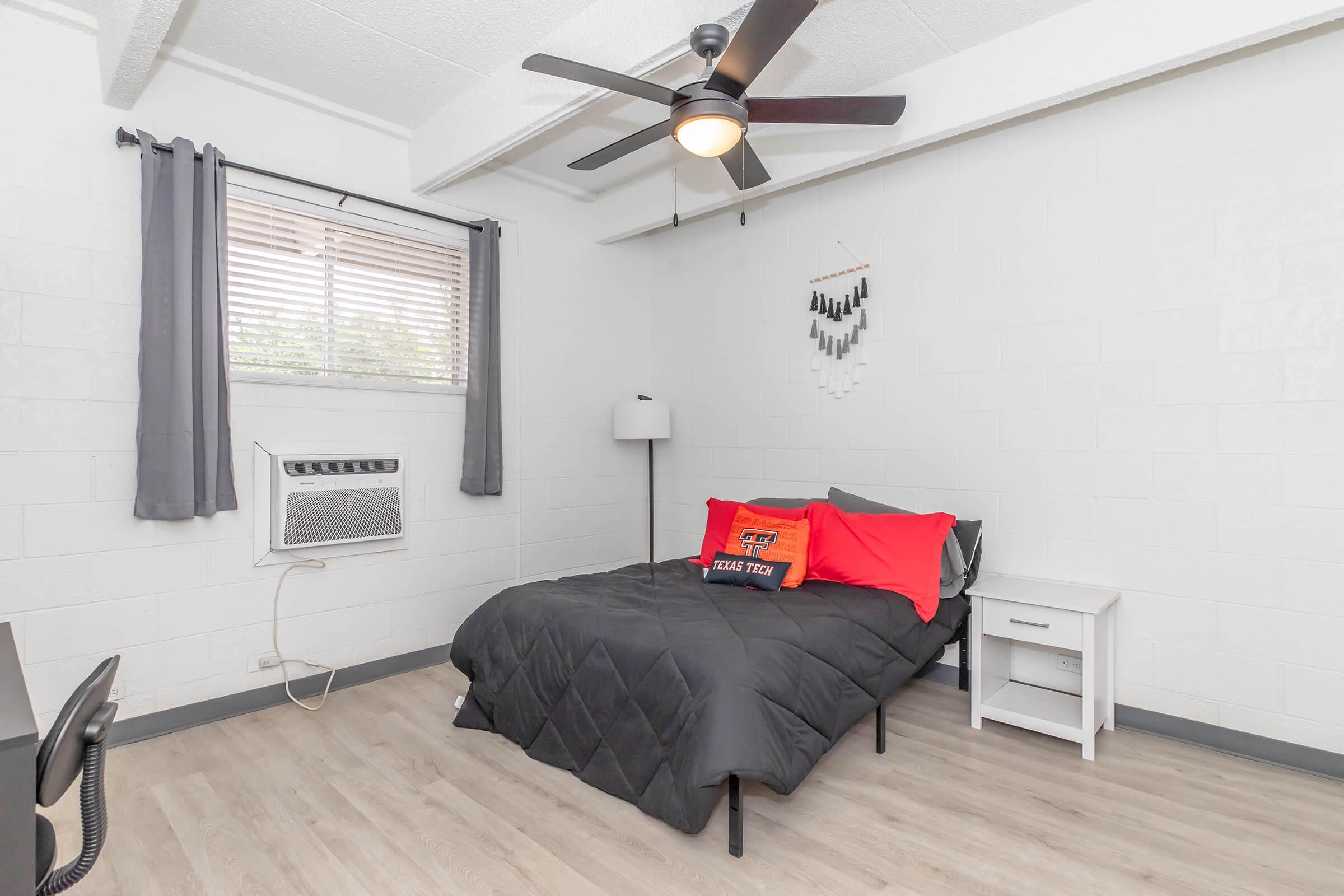 Cozy bedroom featuring a black bedspread with red accent pillows, a bedside table, a standing lamp, and an air conditioning unit. Natural light comes through a window with gray curtains, and there's a decorative wall hanging. The floor is light-colored, enhancing the room's spacious feel.
