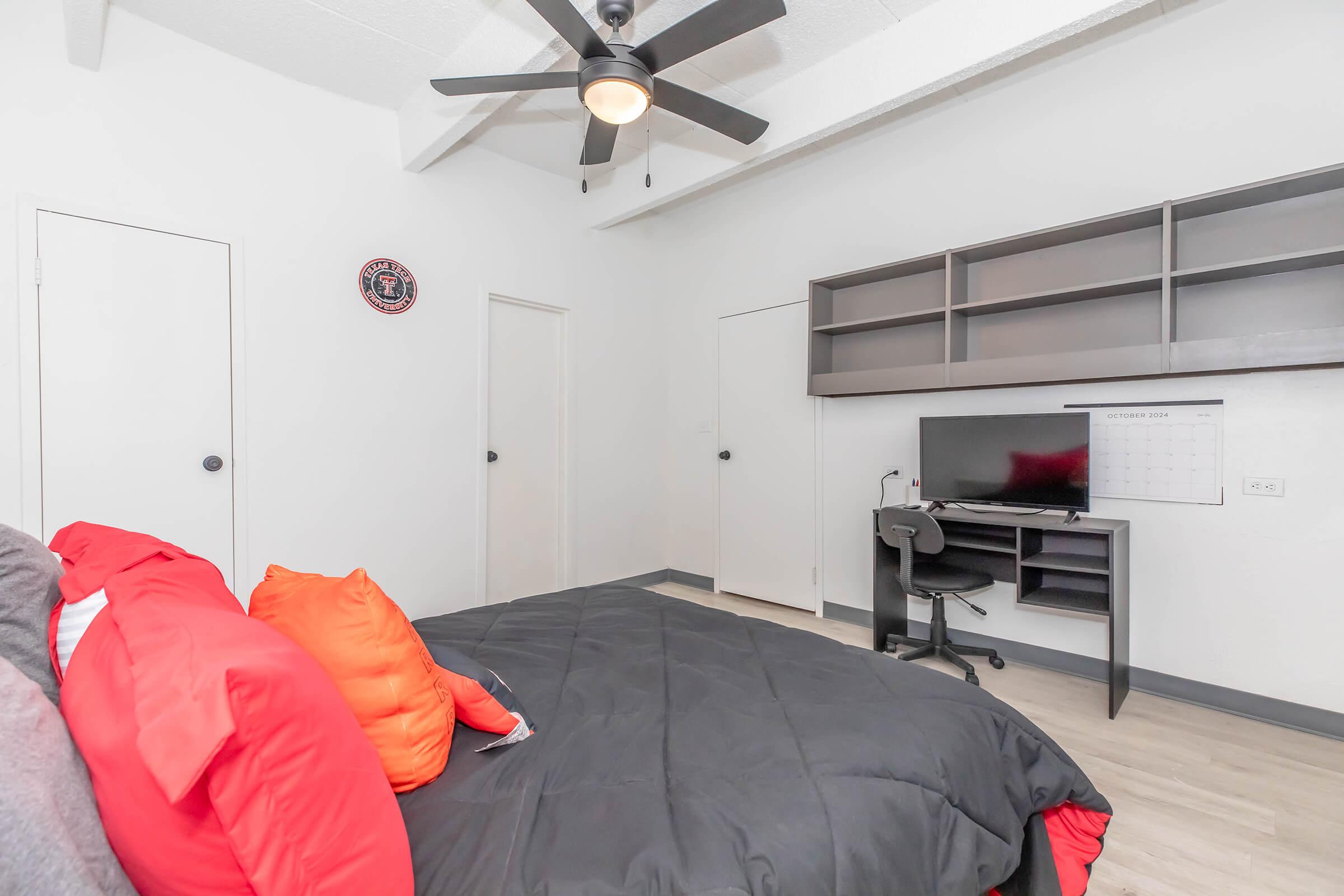 A modern bedroom featuring a black comforter on a bed with orange pillows, wooden flooring, a desk with a chair and computer, a calendar on the wall, and two closed doors. A ceiling fan is visible above, and a wall-mounted decoration can be seen. The room has a minimalist, clean design.