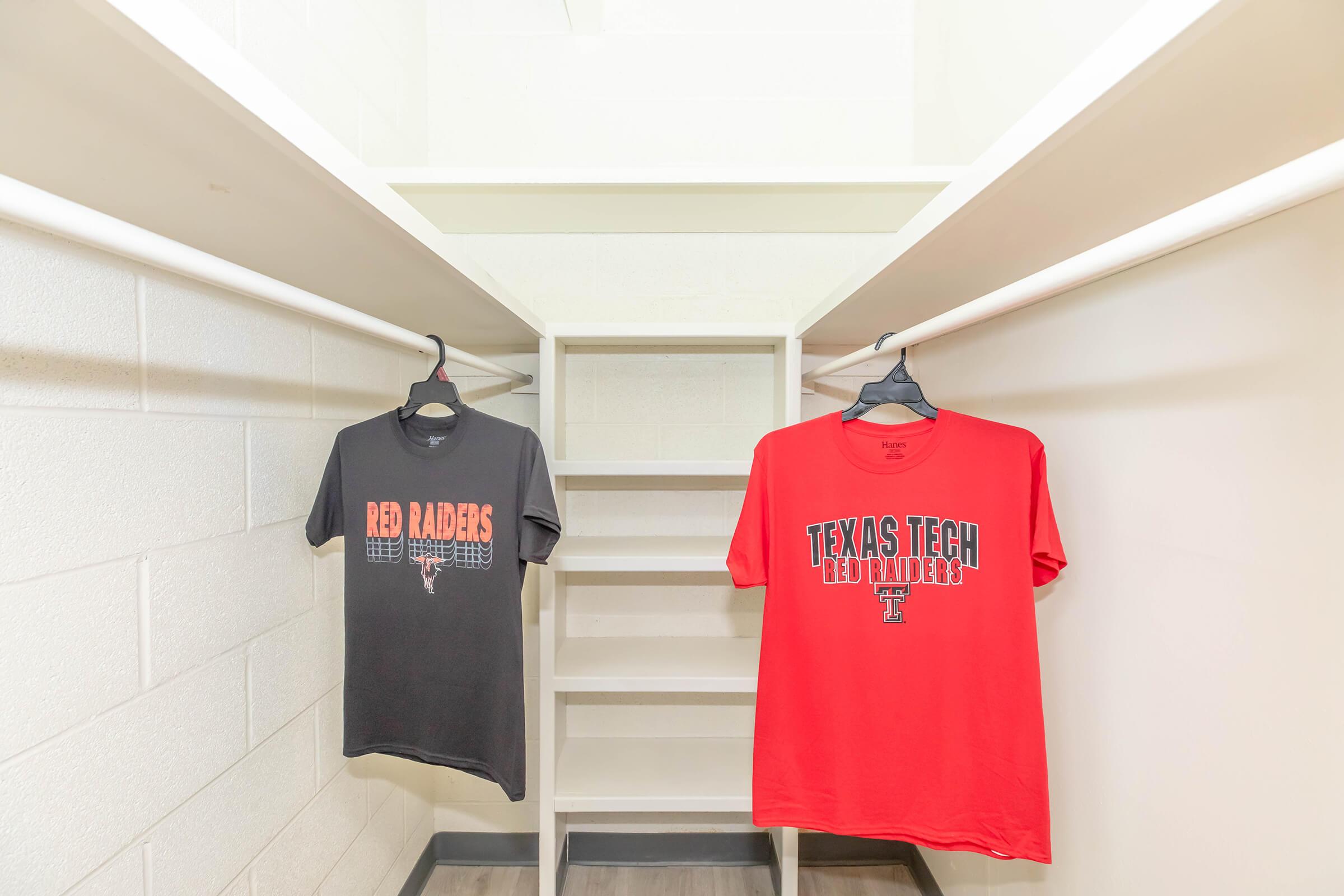 A walk-in closet featuring two t-shirts on hangers. One shirt is black with "Red Raiders" printed on it, and the other is red with "Texas Tech Red Raiders" displayed prominently. The closet has white walls and a shelf unit in the background.