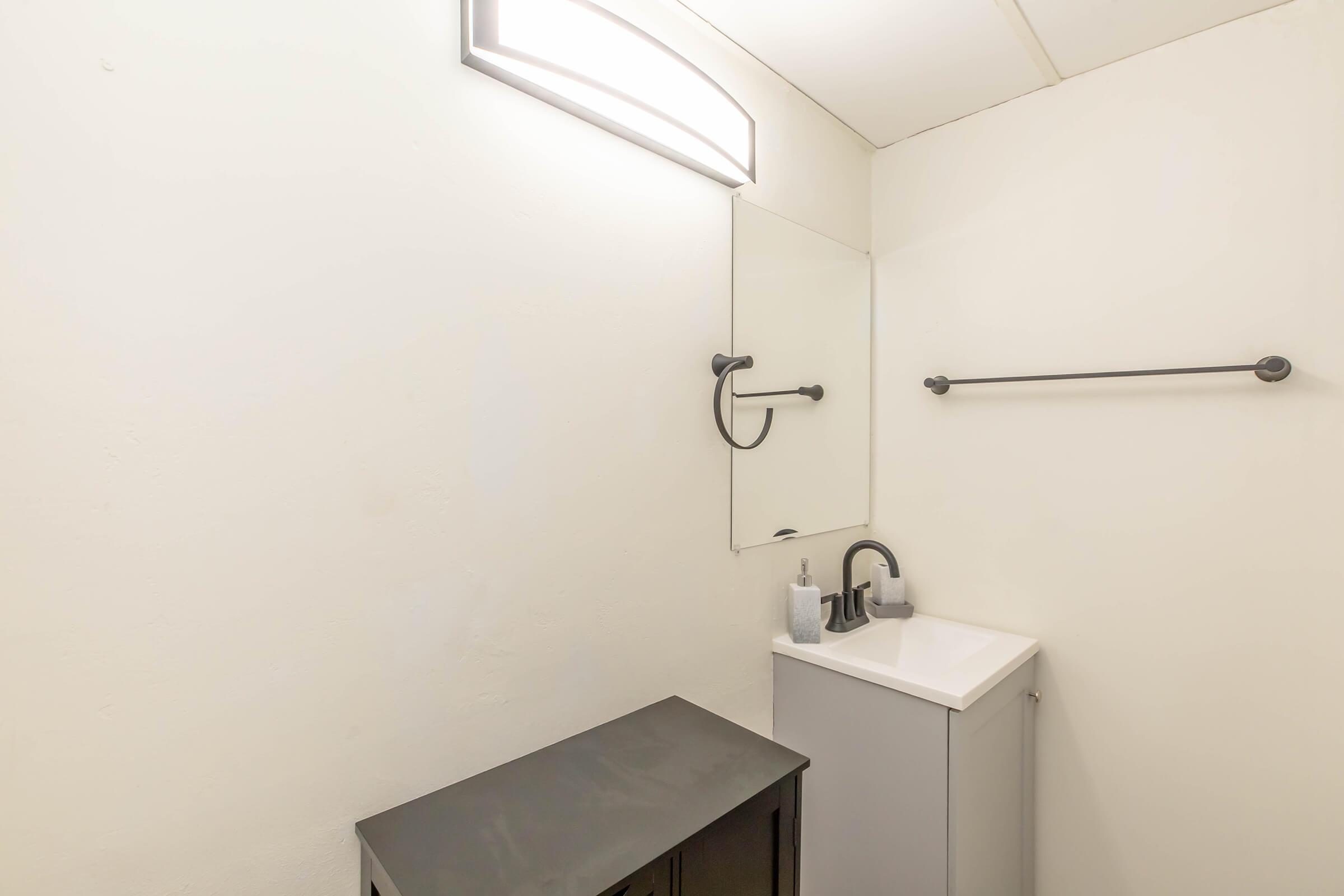 A simple bathroom interior featuring a white wall, a small vanity with a sink and faucet, a mirror above the sink, and a towel bar on the right. There's a dark cabinet or shelf below the sink, and a light fixture above the mirror providing illumination.