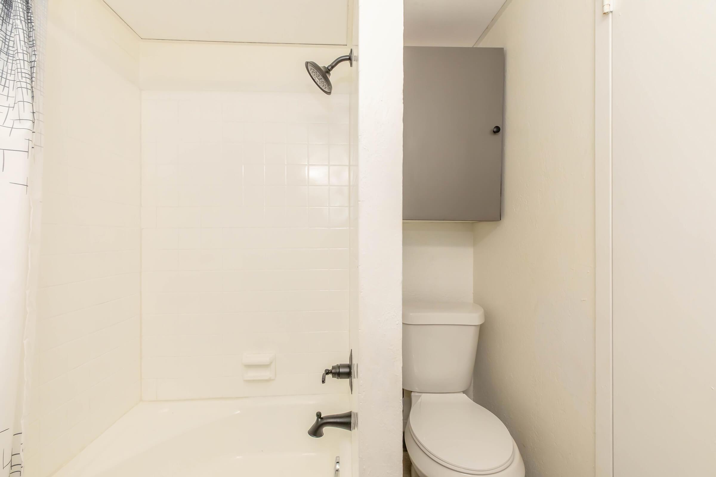 A bathroom with a shower area on the left, featuring white tiled walls and a showerhead. On the right, there is a closed toilet next to a small cabinet mounted on the wall. The overall design is simple and functional, with neutral colors.