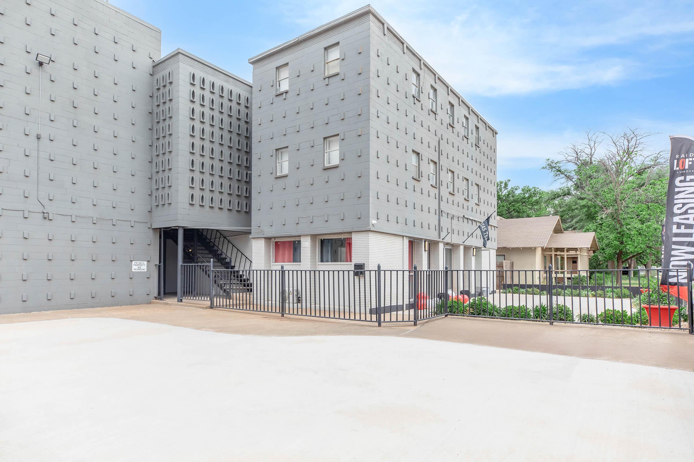 A modern, multi-story apartment building with a gray exterior featuring a distinctive pattern. The building has a gated entrance, a staircase leading to upper floors, and a landscaped area with plants. In the foreground, there’s a clear, paved space. A leasing sign is visible on the right.
