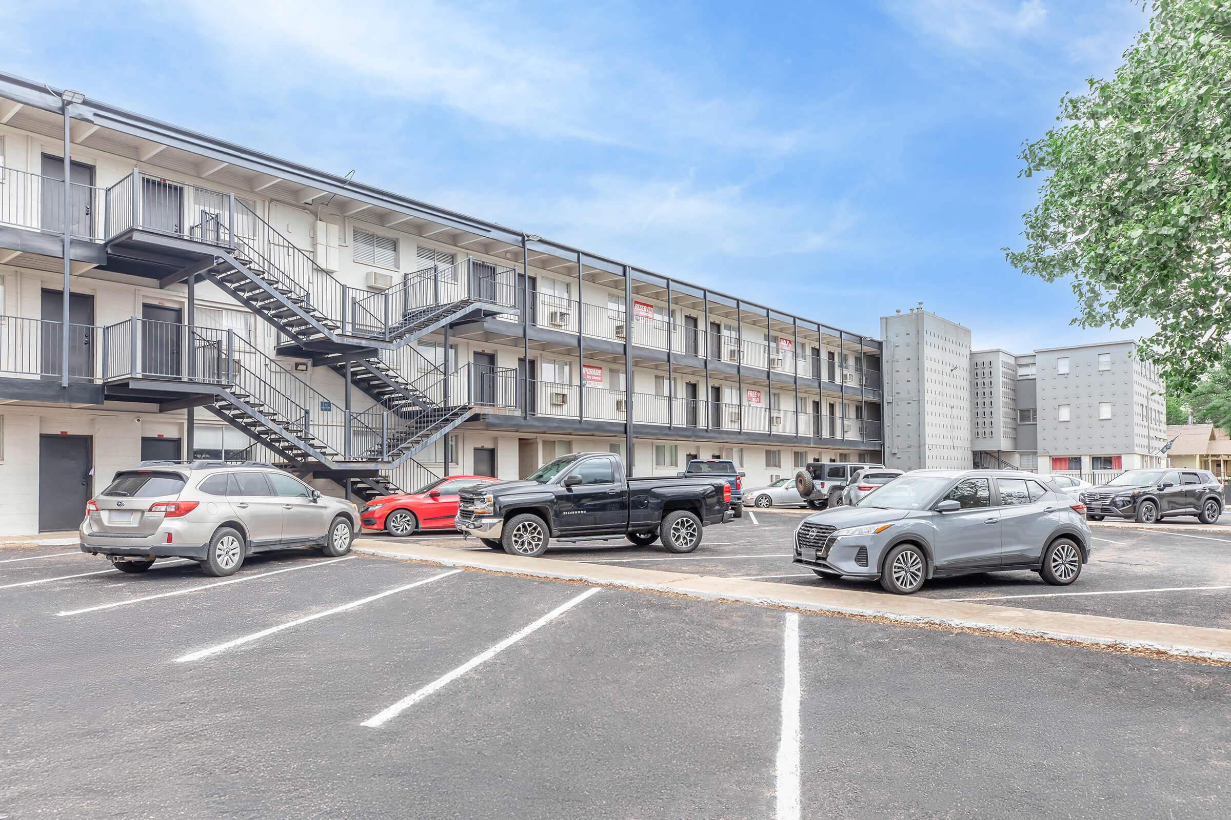 Exterior view of a two-story motel with a black metal staircase. Several cars are parked in front, including a black pickup truck, a silver SUV, and a red car. The building features multiple rooms with balconies, and a blue sky is visible in the background.