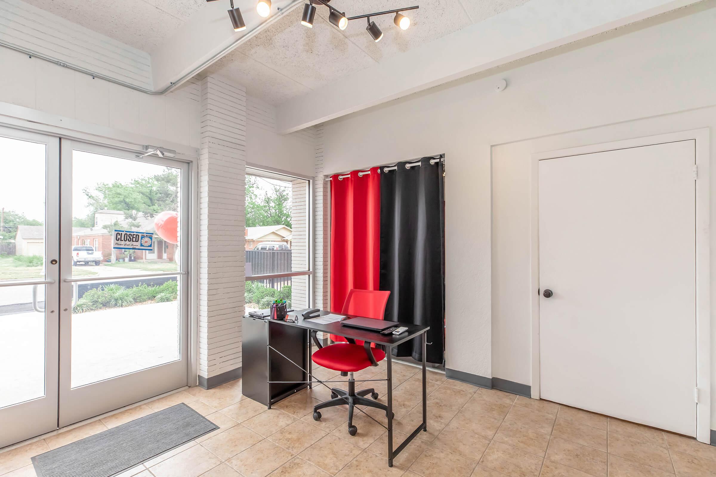Interior of a modern office space featuring a black desk with a red chair, situated near large glass doors. The walls are light-colored, and there are black and red curtains. A door leads to another room, and a welcome mat is placed near the entrance. Natural light illuminates the area, enhancing a professional atmosphere.