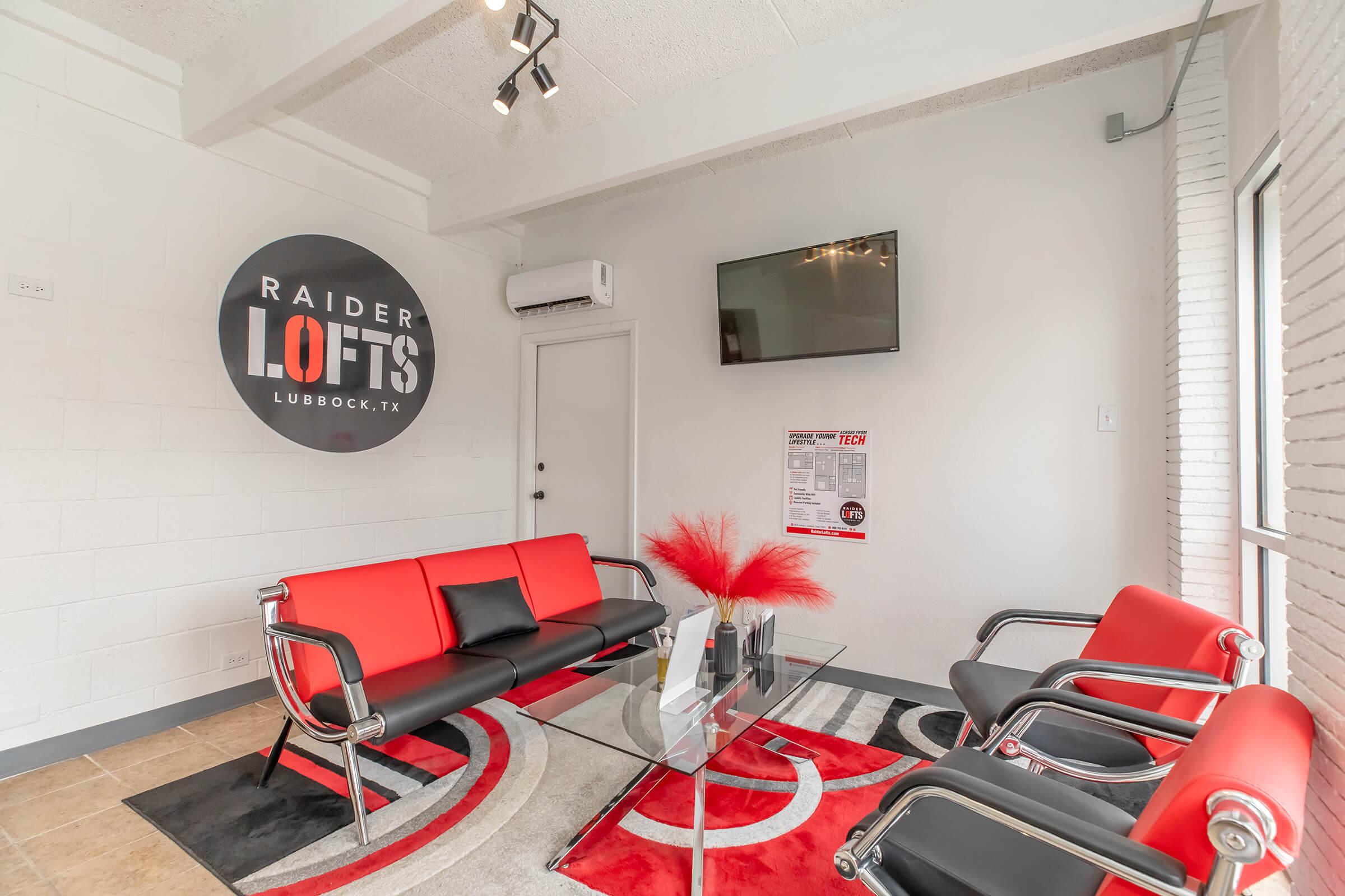 A modern lounge area featuring sleek black and red furniture, including a couch and chairs, set on a stylish rug. The wall displays a circular sign for "Raider Lofts" in Lubbock, Texas. A flat-screen TV is mounted, and the room is well-lit with natural light.