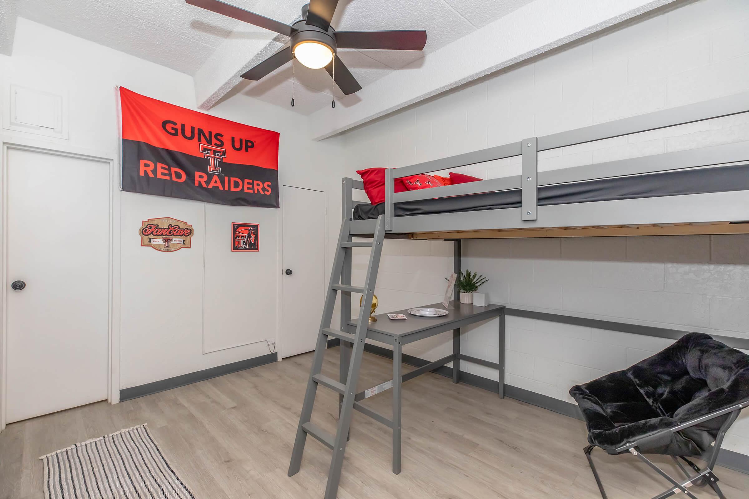 A student bedroom featuring a lofted bed with a ladder, a wall adorned with a Texas Tech University "Guns Up Red Raiders" banner, a small desk with a plant, and a black floor chair. The room has light-colored walls and wooden flooring, creating a cozy and functional study space.