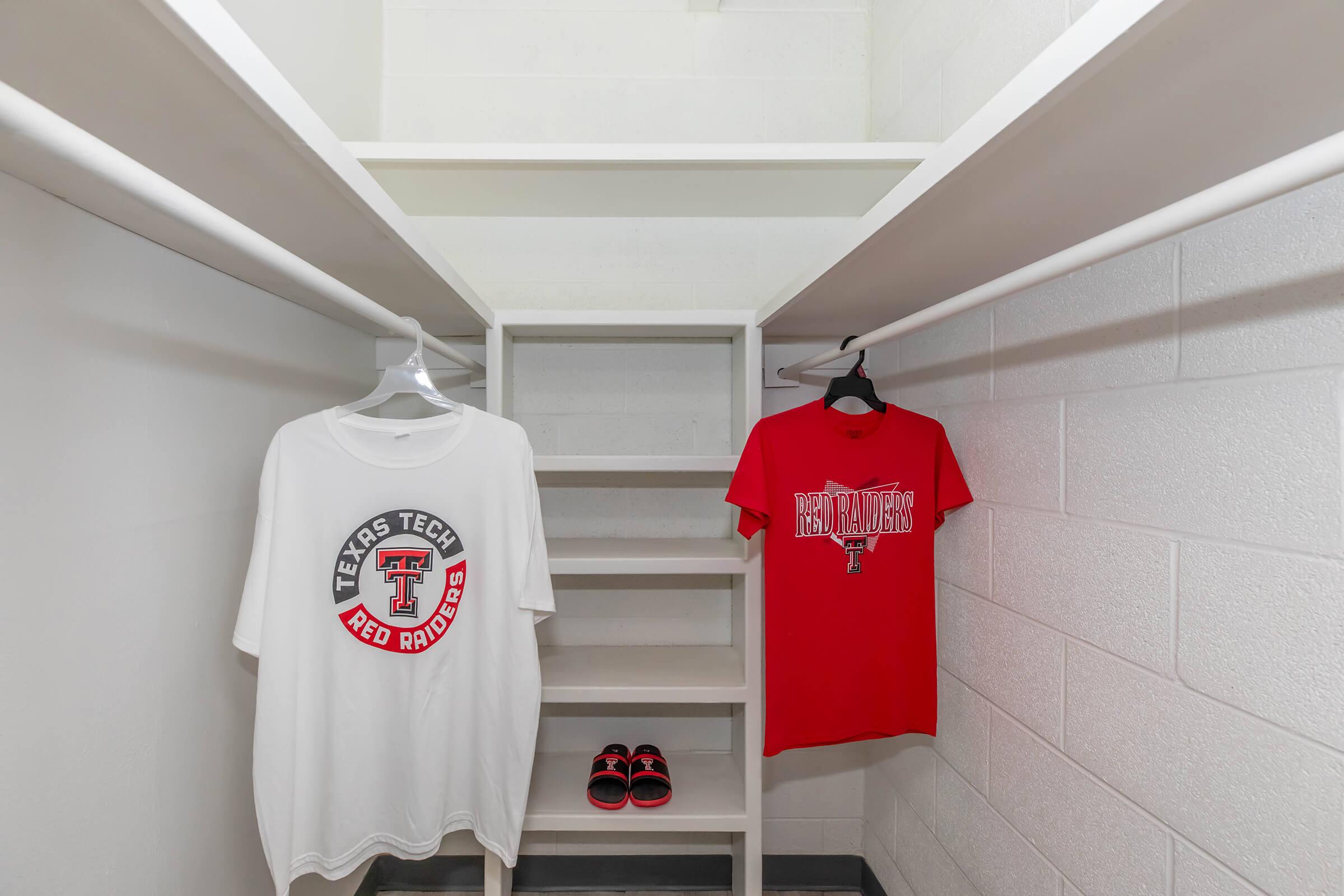 A well-organized closet with two Texas Tech Red Raiders T-shirts hanging on hangers—one white with a logo and the other red. There are several shelves in the background, and a pair of red sandals is placed on the floor. The walls are painted white, creating a clean, minimalist look.