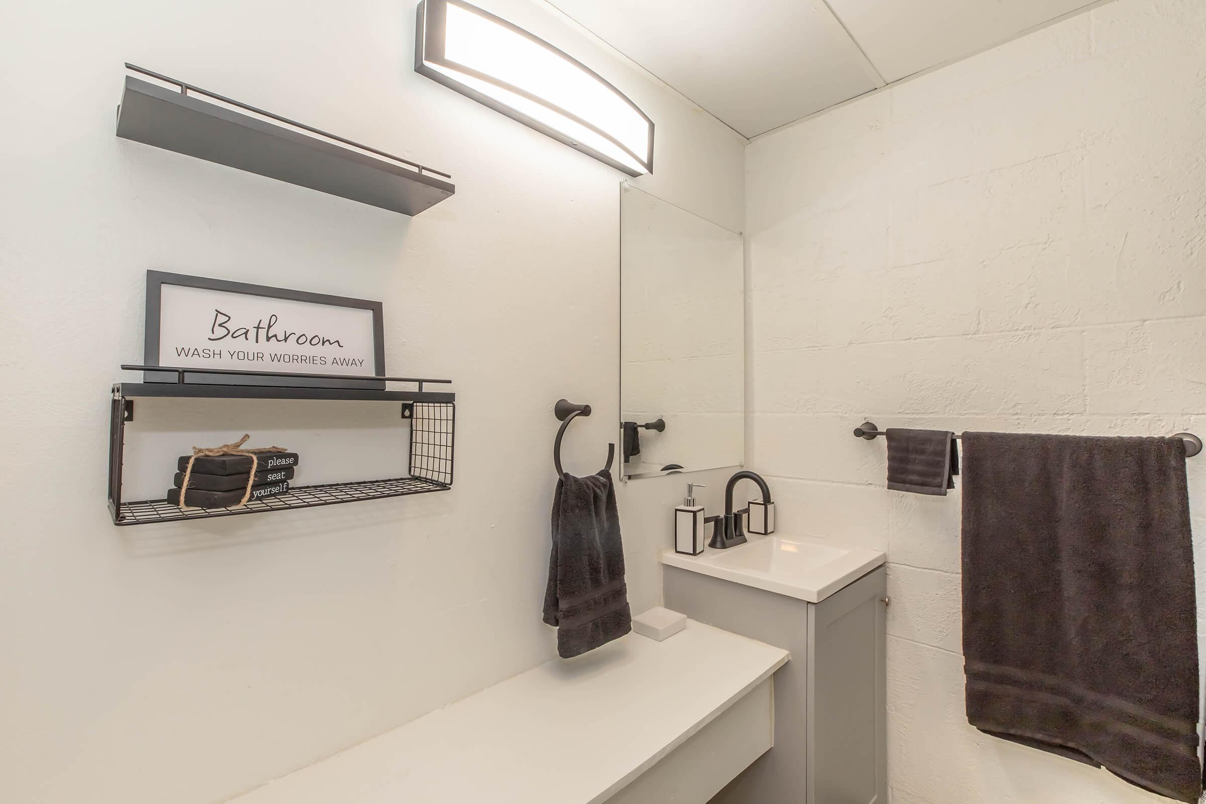 A modern bathroom featuring a wall-mounted mirror, a sleek sink with a faucet, and a small shelf holding decor items. Black towels hang on a rack, with a light fixture above. A sign on the shelf reads "Bathroom - WASH YOUR WORRIES AWAY," adding a decorative touch to the minimalist design.