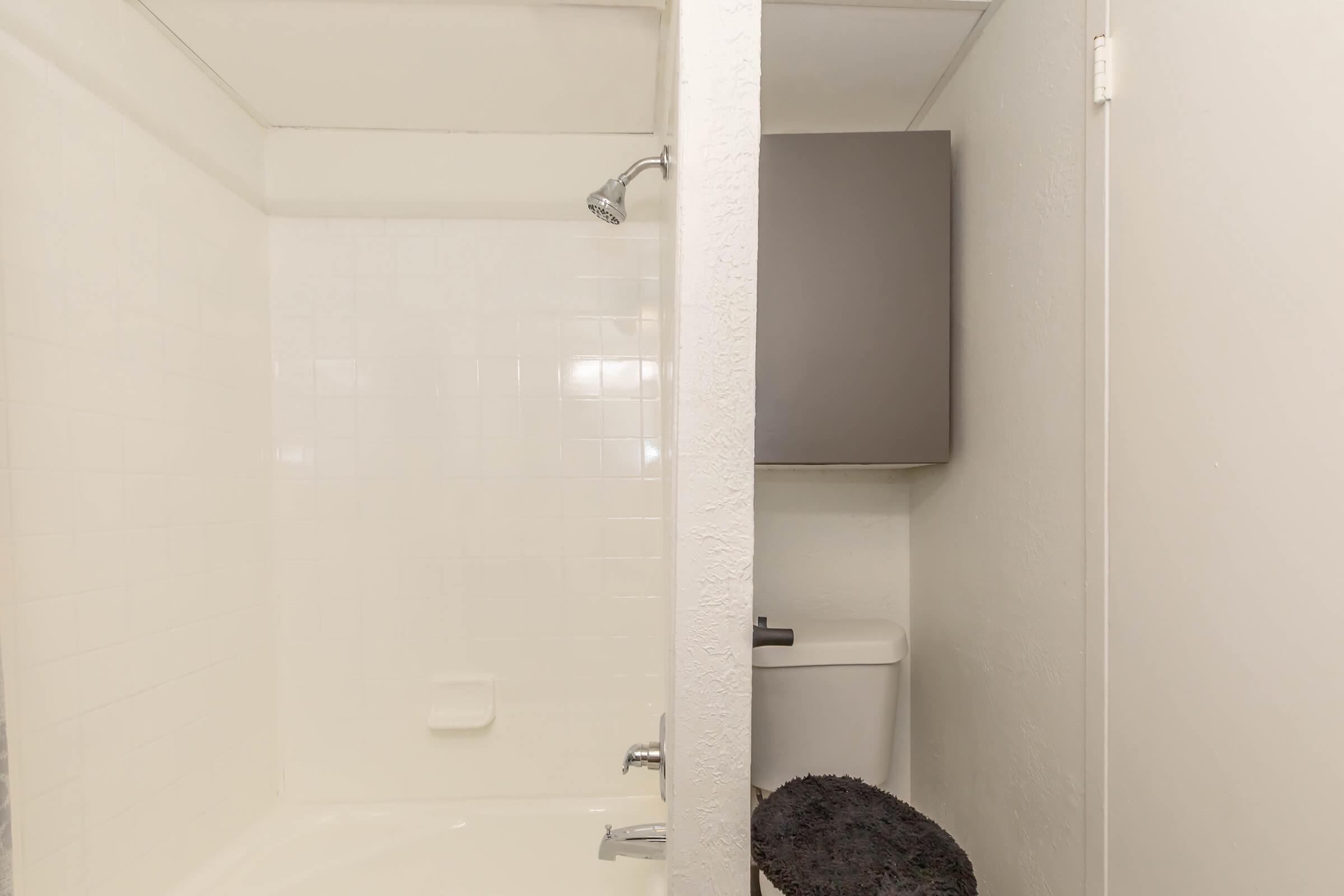 Modern bathroom featuring a white tiled shower with a silver showerhead on the left. On the right, there's a toilet partially obscured by a wall, with a dark rug in front. The overall design is simple and clean with neutral colors.
