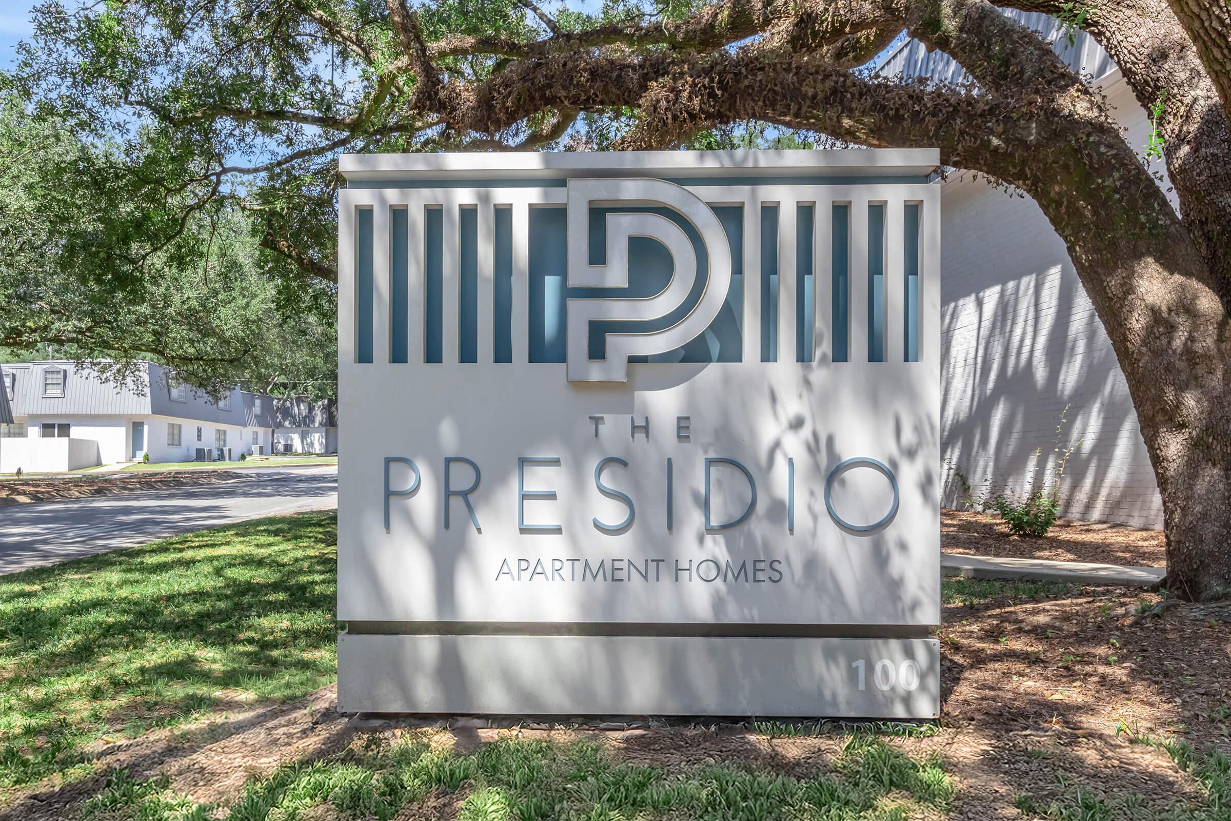 Sign for The Presidio Apartment Homes, featuring a modern design with the initials "P" integrated into the logo. The sign is situated outdoors under a tree, with the address "1106" visible. The surrounding area includes green grass and landscaping.