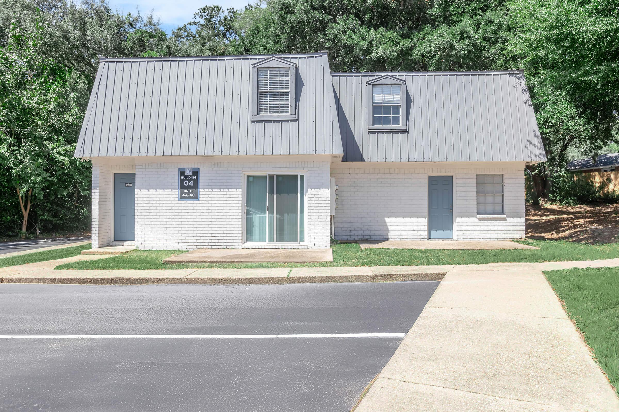 A single-story residential building with a gray metal roof and white brick exterior. It features two separate units, each with a door and window, situated on a landscaped lot with trees in the background. A concrete walkway leads to the front entrance, and a paved road runs alongside the property.