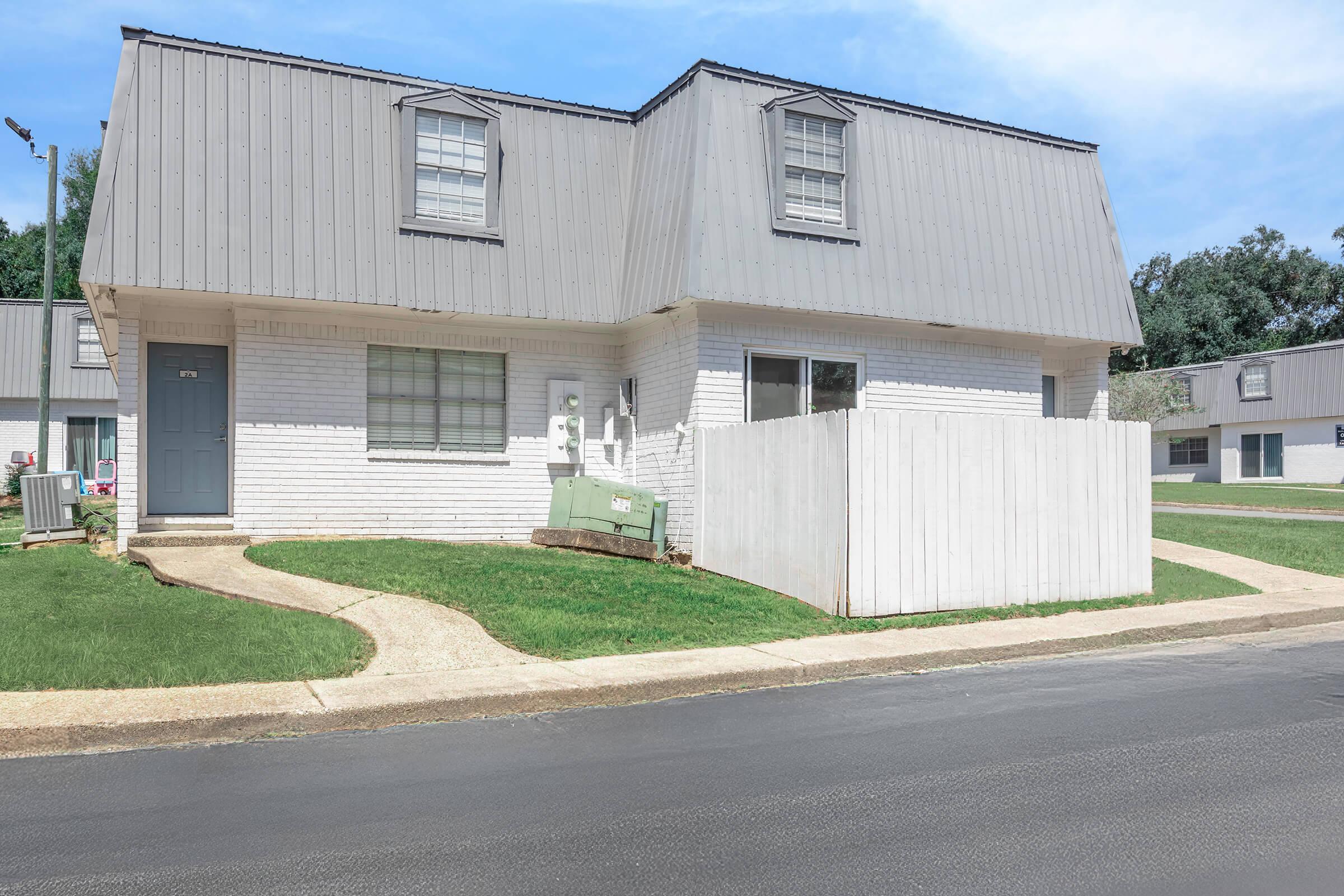 A two-story white brick duplex with a modern metal roof, featuring a small front lawn and a pathway leading to the entrance. A white fence partially encloses the yard, with a mailbox visible. Trees and additional buildings can be seen in the background under a clear blue sky.