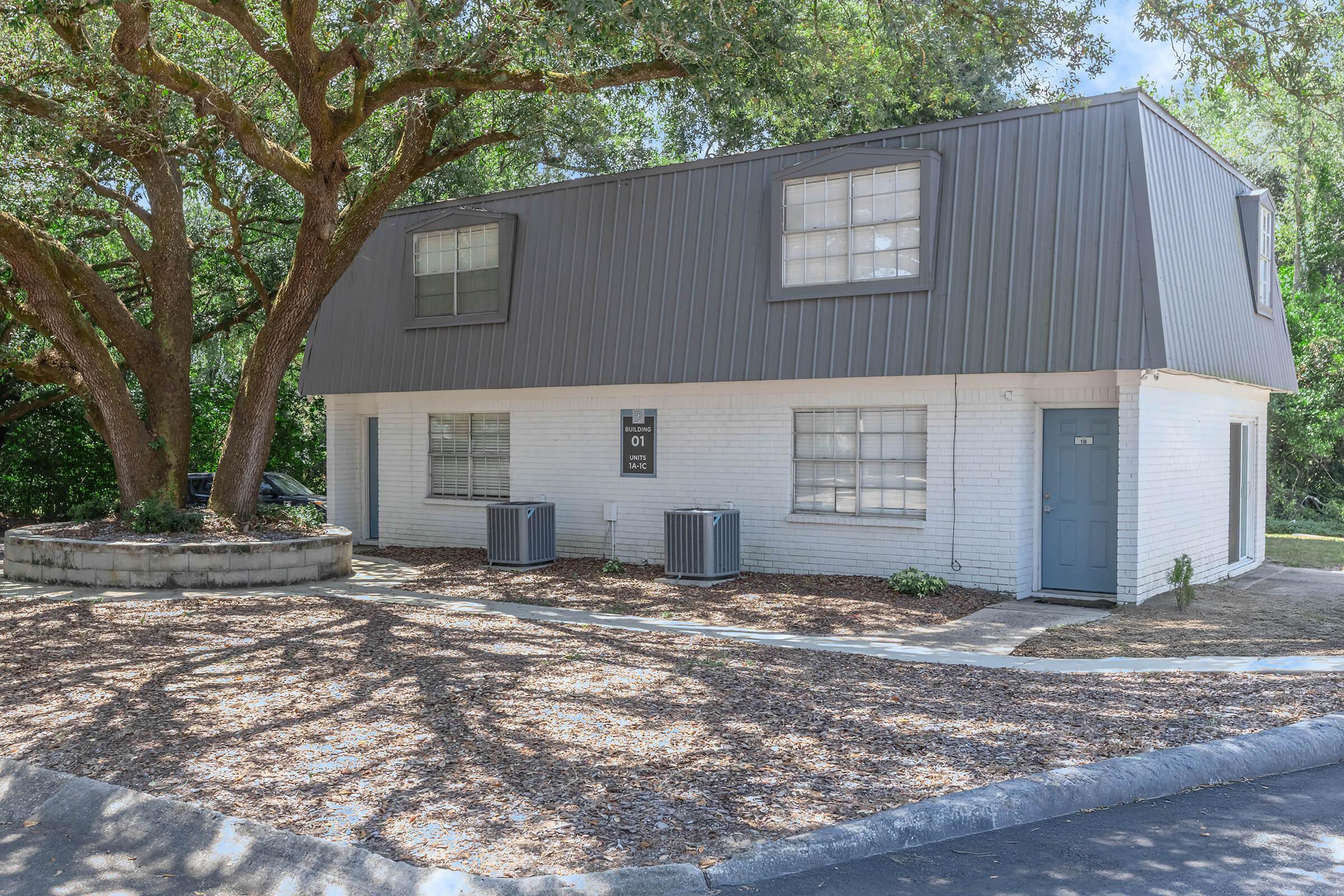 Two-story building with a modern design, featuring a gray metal roof and white brick walls. The front has two air conditioning units and several windows. Surrounding the building is landscaped ground cover with mulch and a large tree providing shade. The entrance is marked by a blue door.