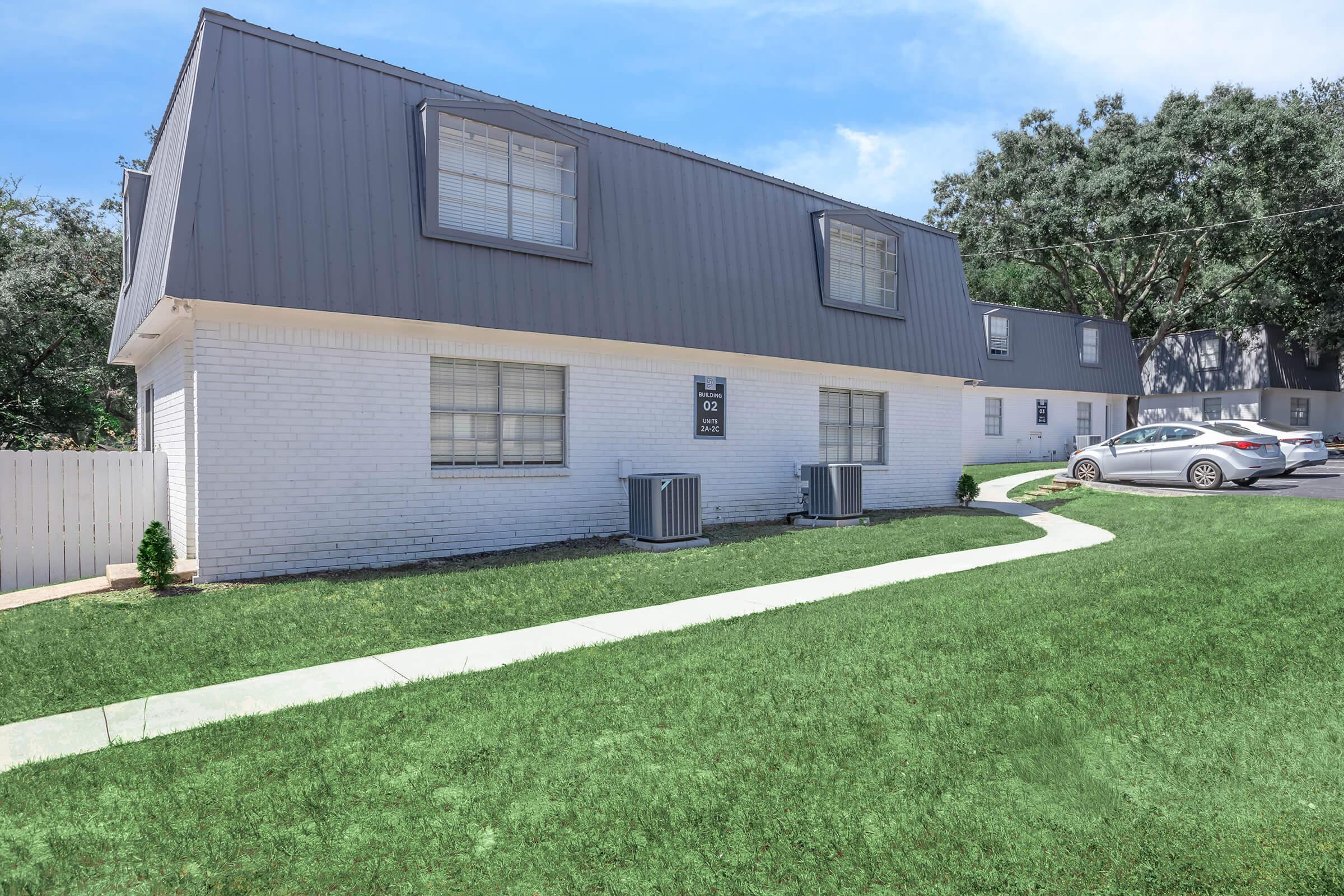 A two-story residential building with a gray and white exterior, featuring multiple windows. The front yard is grassy with a curved walkway leading to the entrance. Two air conditioning units are visible on the ground, and a gray car is parked nearby. Trees are visible in the background.