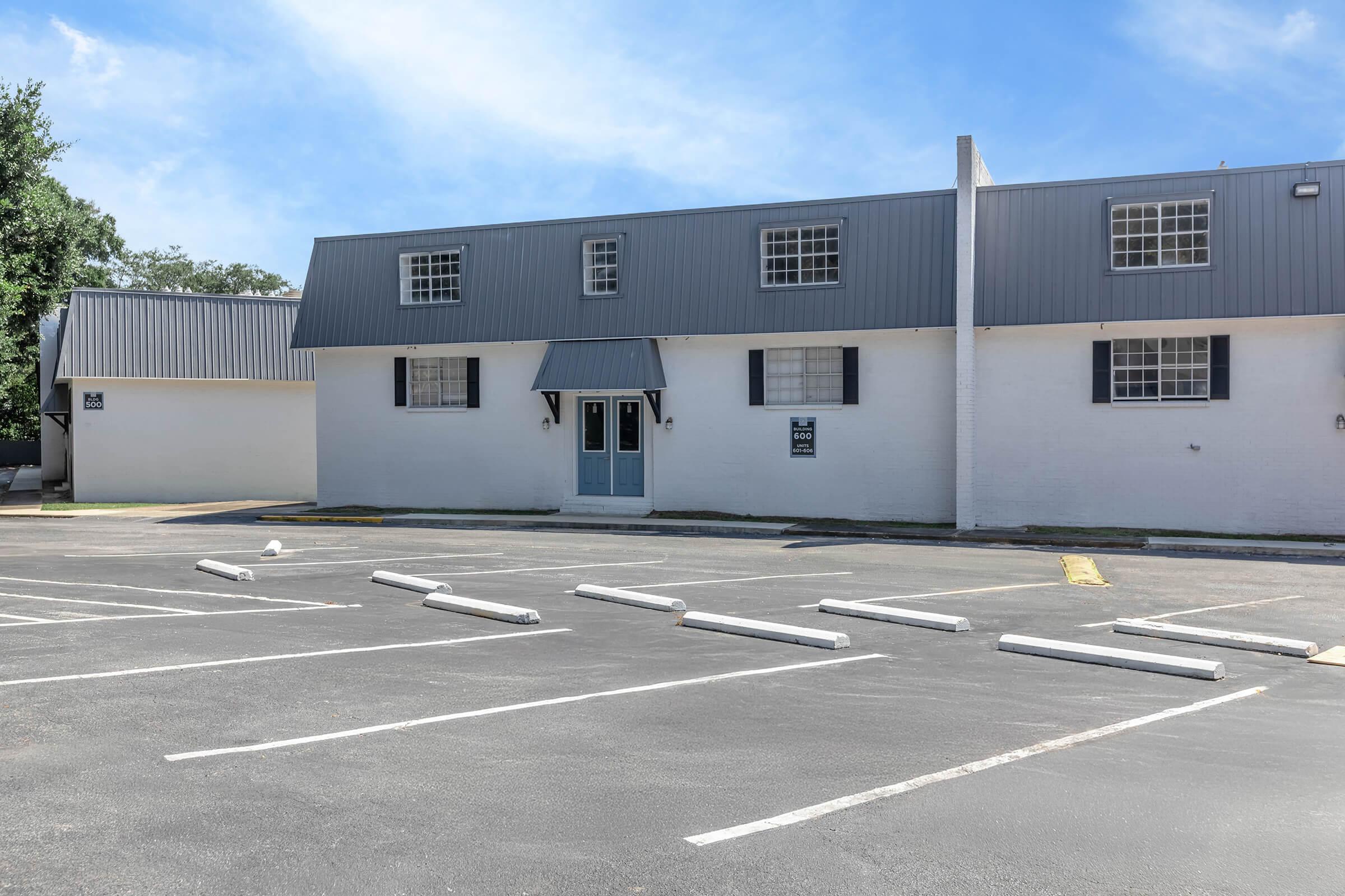 A view of a building featuring a gray roof and white walls, with multiple windows and a blue front door. An empty parking lot with marked spaces is in the foreground, and clear blue sky can be seen in the background.