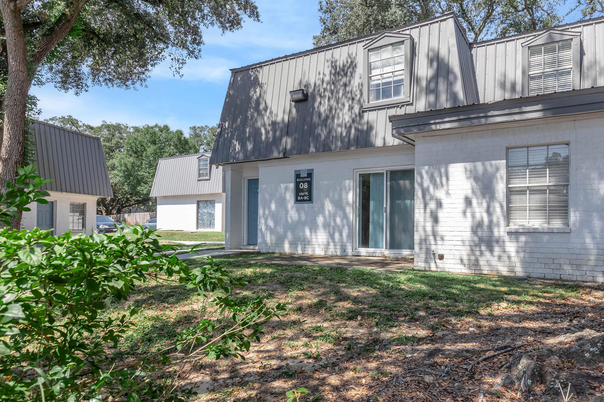 A view of a residential building with a light-colored brick exterior and gray metal roof. The building features several windows and a front door. In the foreground, there is green foliage and well-maintained grass. The scene is set during the day with clear blue skies.