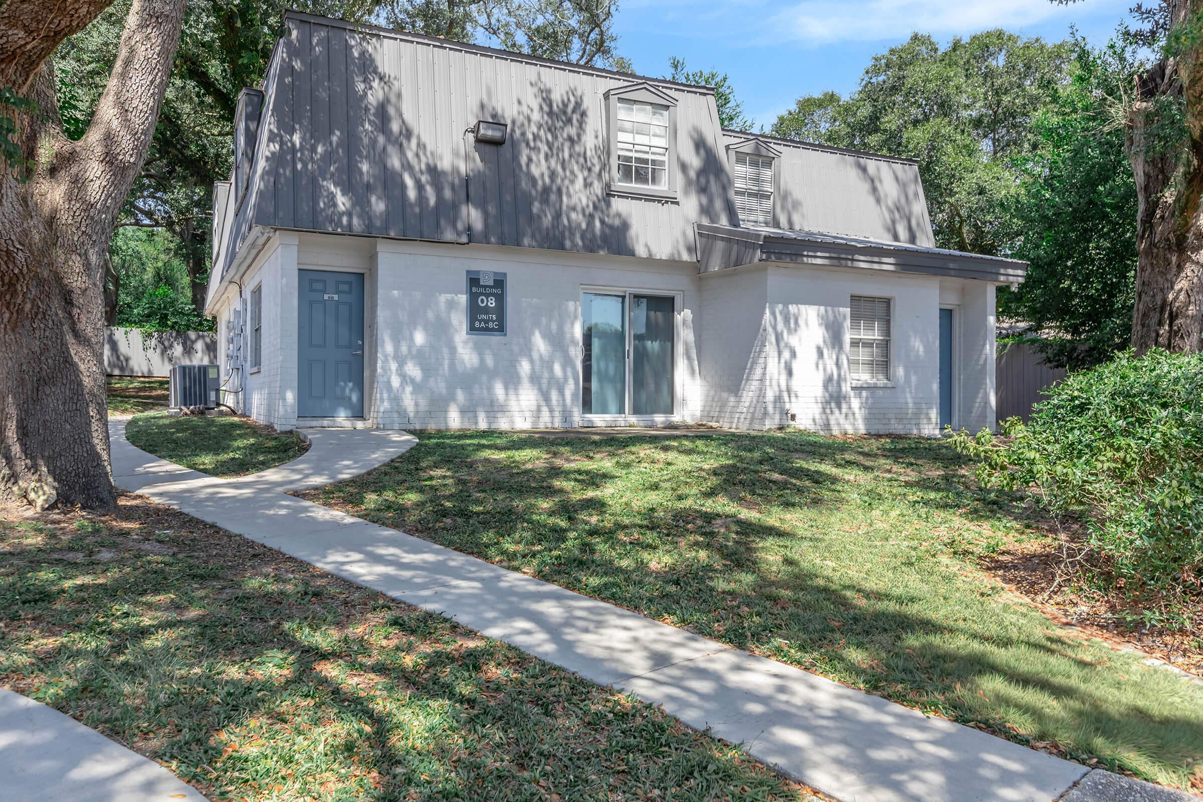 Two-story house with a gray roof and white exterior, featuring blue front doors and several windows. The house is surrounded by a well-maintained lawn and a walkway leading to the entrance, with trees providing shade. Bright, sunny day with clear blue skies.