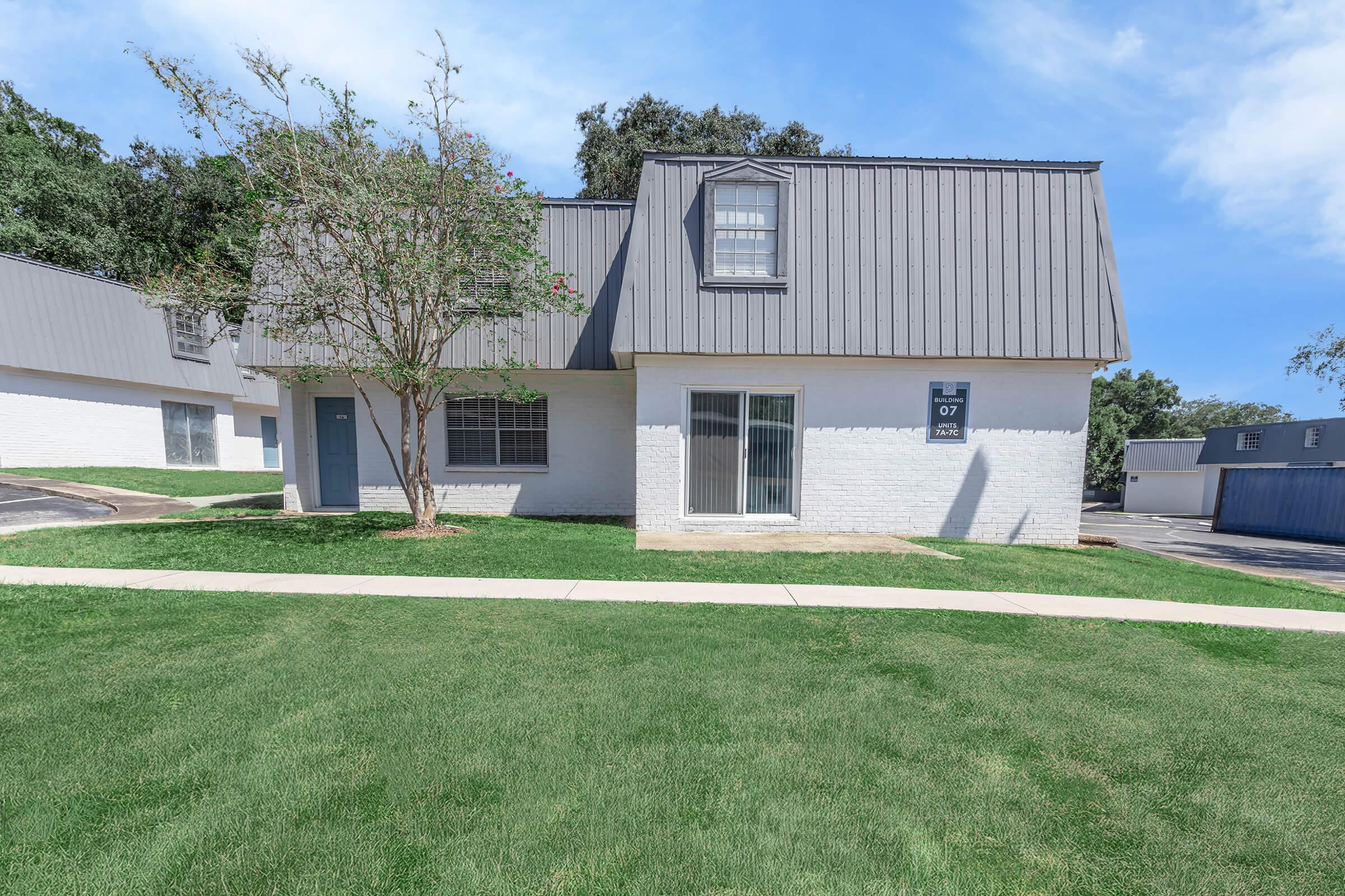 A two-story residential building with a modern, sloped roof. The exterior features white walls with gray accents. A small tree is planted in front, and there is a pathway leading to the entrance. The scene has a clear blue sky in the background, highlighting a well-maintained lawn and parking area.
