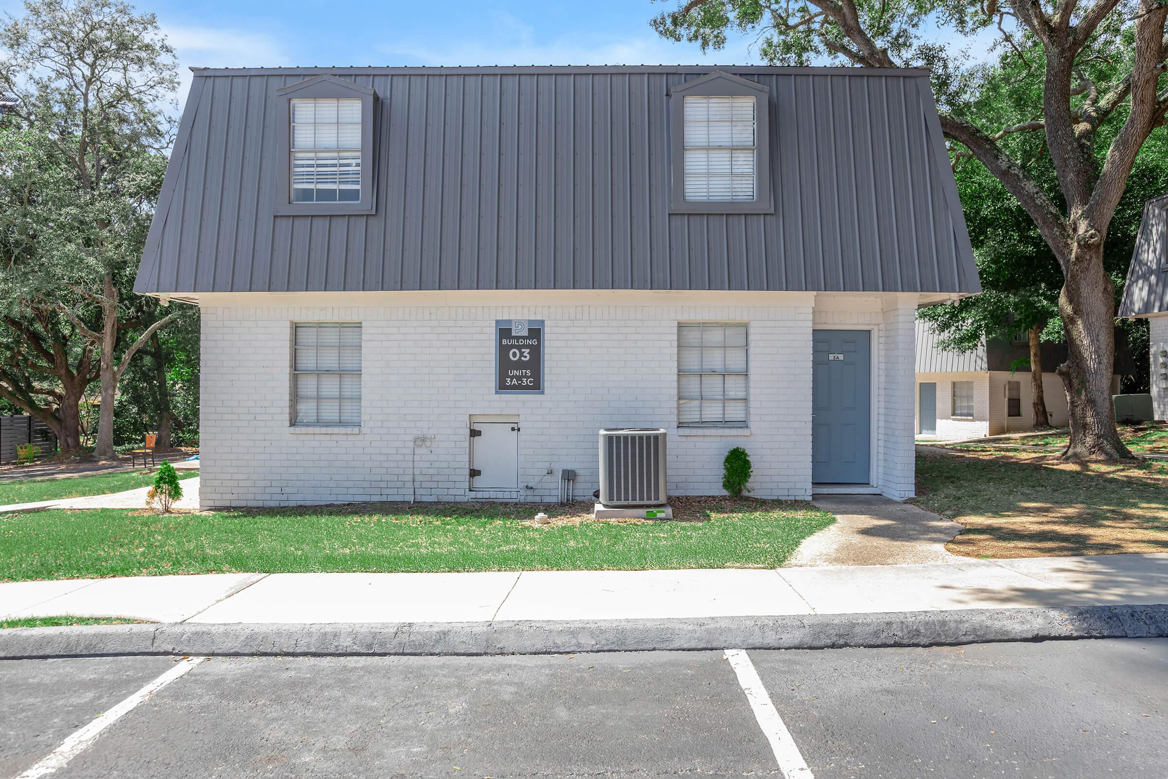 Two-story building with a sloped gray metal roof and white brick exterior. The front features a blue door, a small air conditioning unit, and a sign displaying "03" near the entrance. Surrounding the building is well-kept greenery, sidewalks, and a parking area. Trees are visible in the background.