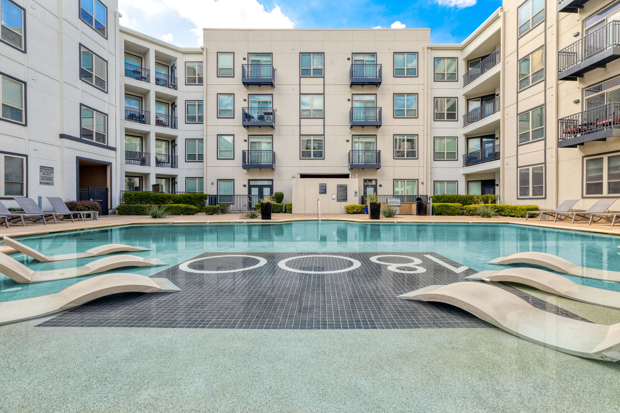 A modern apartment complex with a clear blue pool at the center. The pool features sleek lounge chairs along the edges, and the background shows multiple floors of contemporary apartments with balconies. The area is well-maintained, with greenery around the poolside.