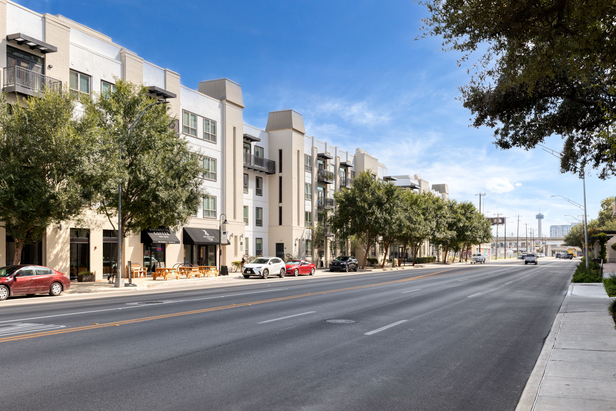 A quiet urban street lined with modern apartment buildings and trees. There are parked cars and some outdoor seating at street-level cafes. The sky is clear with a few clouds, and the road appears well-maintained, indicating a vibrant neighborhood.