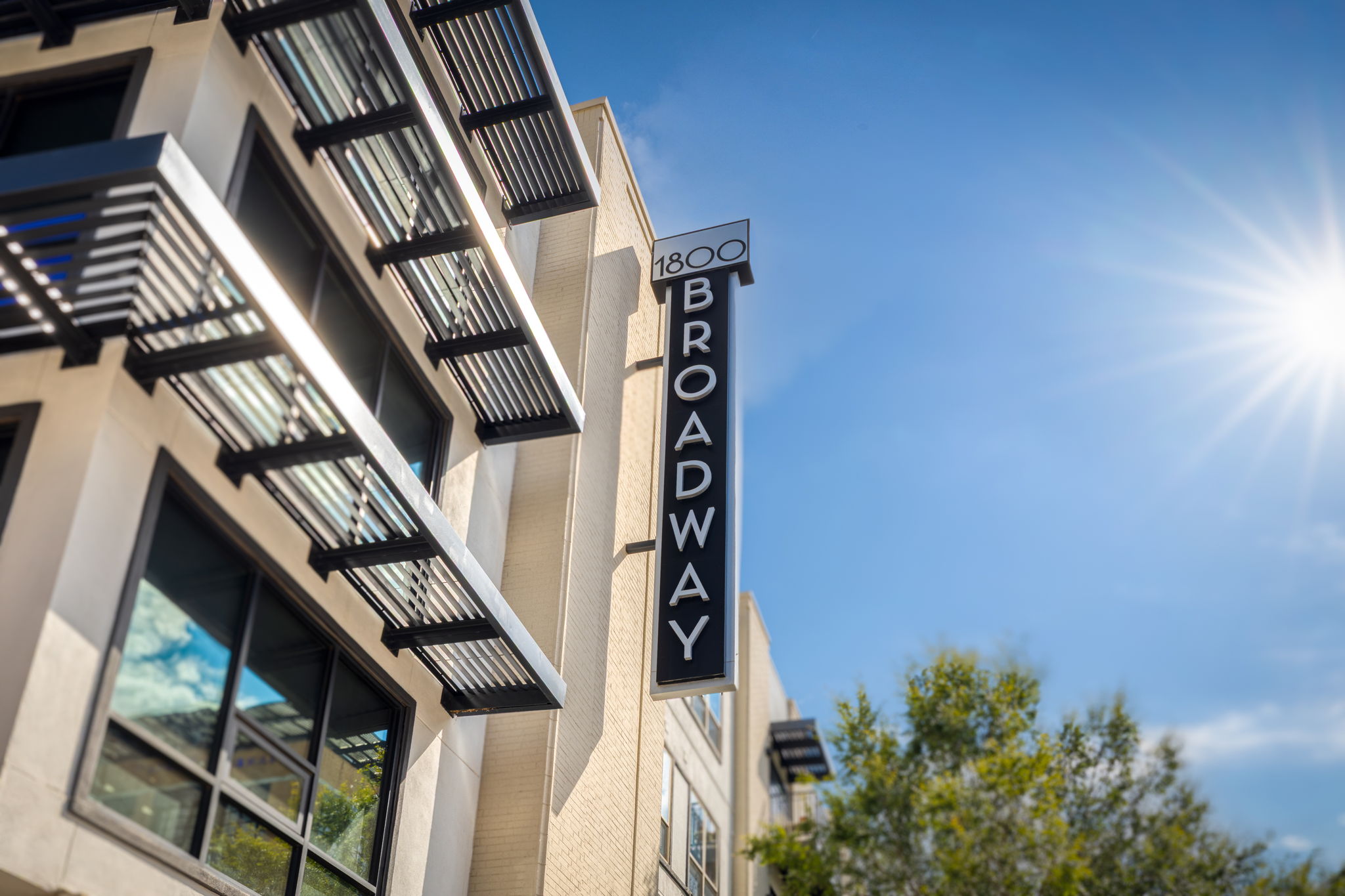 A view of the building exterior featuring the sign "1800 BROADWAY." The facade includes modern architectural elements with large windows and overhanging balconies, set against a clear blue sky with sun rays shining. Green trees are visible in the foreground.