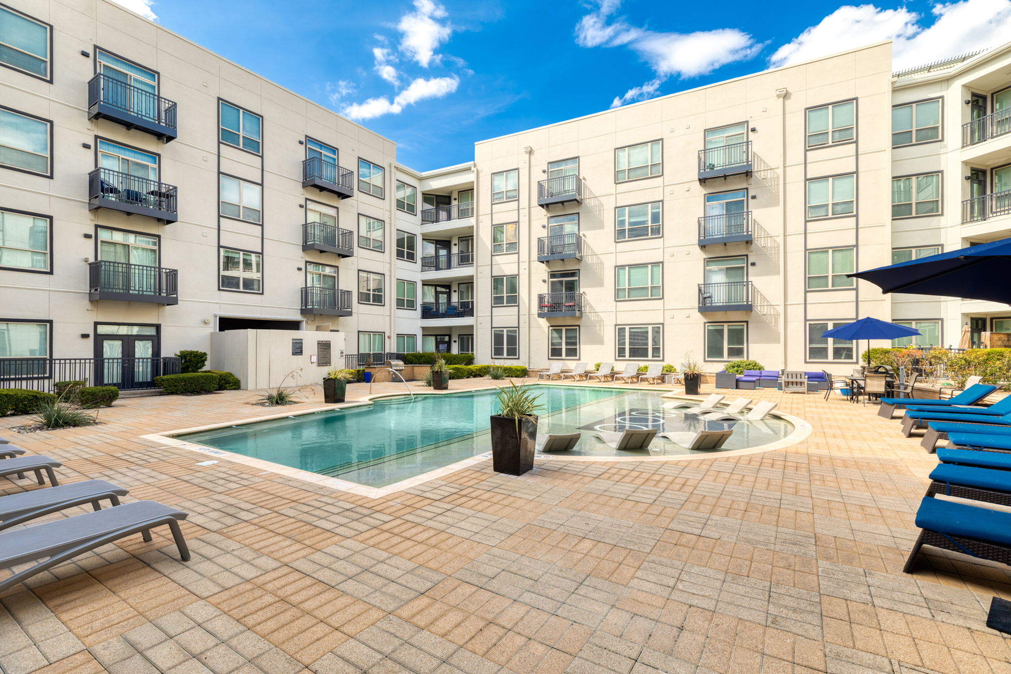A modern apartment complex courtyard featuring a sparkling swimming pool surrounded by lounge chairs. The area is decorated with potted plants, and buildings with large windows provide a bright and inviting atmosphere under a clear blue sky.