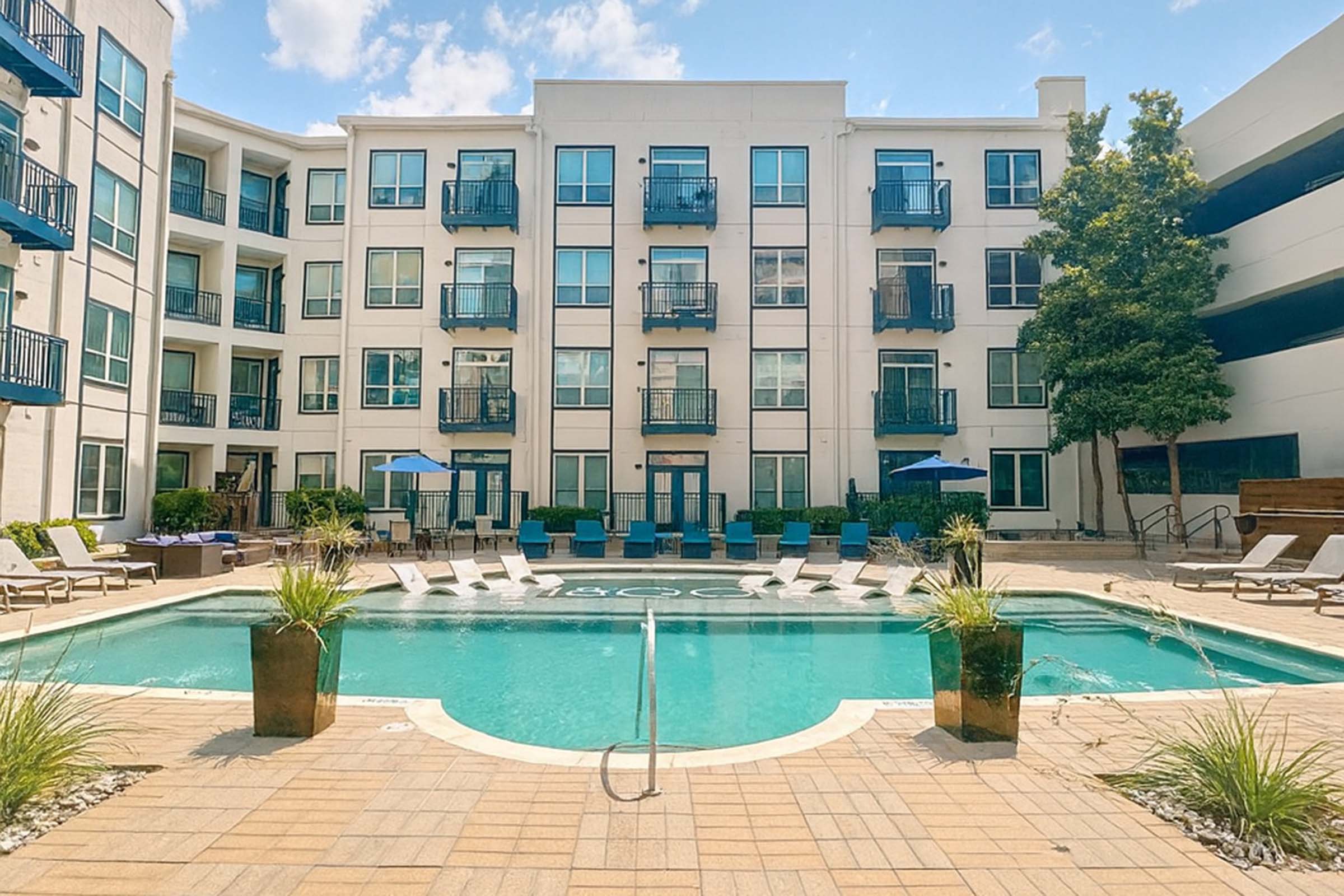 A sparkling outdoor pool surrounded by lounge chairs and potted plants, set in the courtyard of a modern apartment complex. The buildings feature balconies and large windows, with some shaded areas provided by umbrellas. Bright blue skies and a few clouds complete the scene.