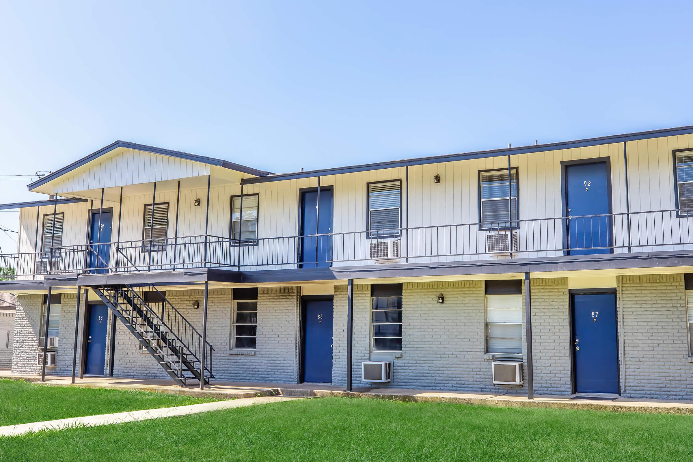 Two-story apartment building featuring a light-colored exterior with blue doors. A metal staircase leads to the upper level, and there are several air conditioning units visible. The grass is neatly maintained, providing a tidy appearance to the property. It's a bright, sunny day with a clear blue sky.