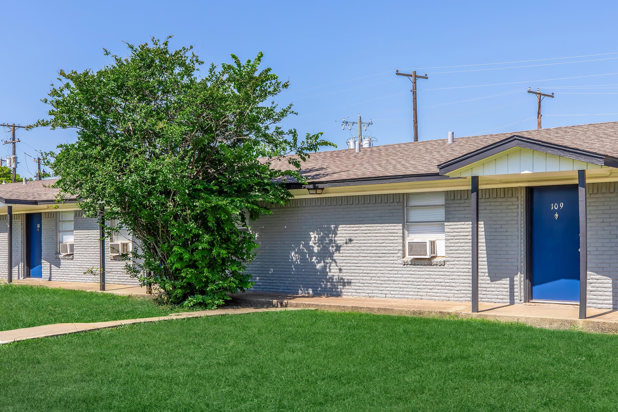 Exterior view of a single-story residential building with several air conditioning units, surrounded by green grass. The building features numbered doors, with a close-up on door number 109. A small tree is positioned near the structure, under a clear blue sky. Utility poles are visible in the background.