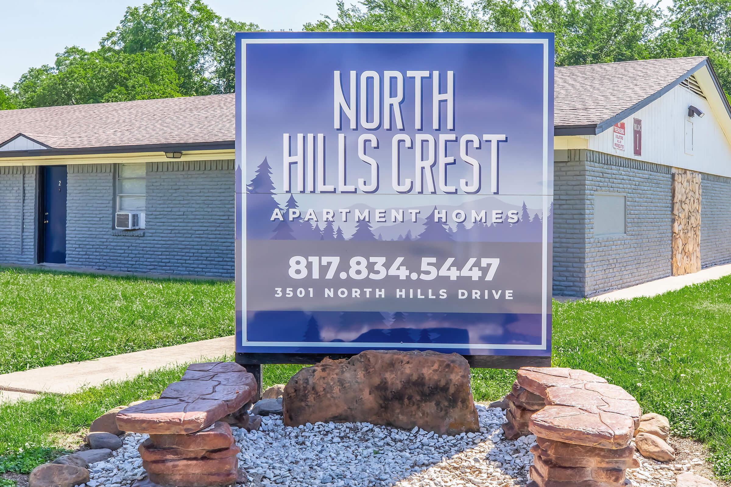 Sign for North Hills Crest Apartment Homes, featuring contact number 817-834-5447 and address 3501 North Hills Drive. The sign is prominently displayed in front of a building, surrounded by a landscaped area with stones and greenery.