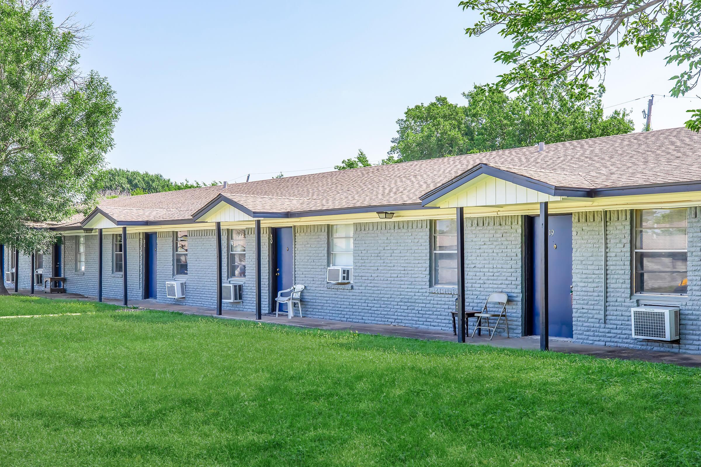 A row of single-story motel rooms with blue doors, situated next to a well-maintained green lawn. Each room features an air conditioning unit and a small chair outside. Trees in the background provide shade, and the clear blue sky adds to the inviting atmosphere.