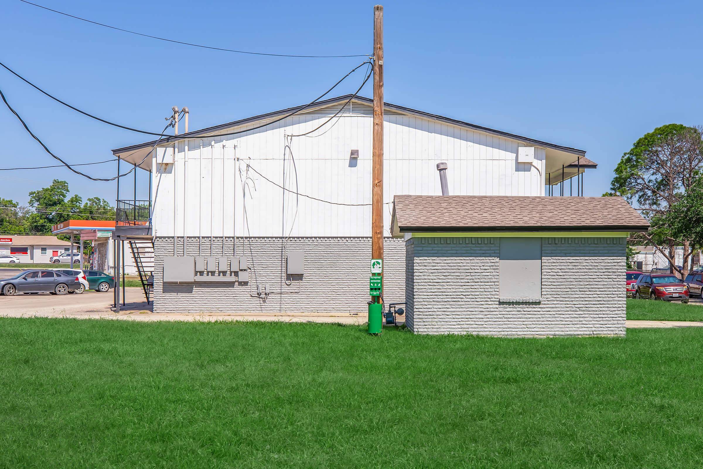 A back view of a simple, single-story building with a white exterior, surrounded by green grass. Visible features include power lines overhead, a small utility structure nearby, and a parking area with some vehicles in the background. The setting appears sunny and well-maintained.