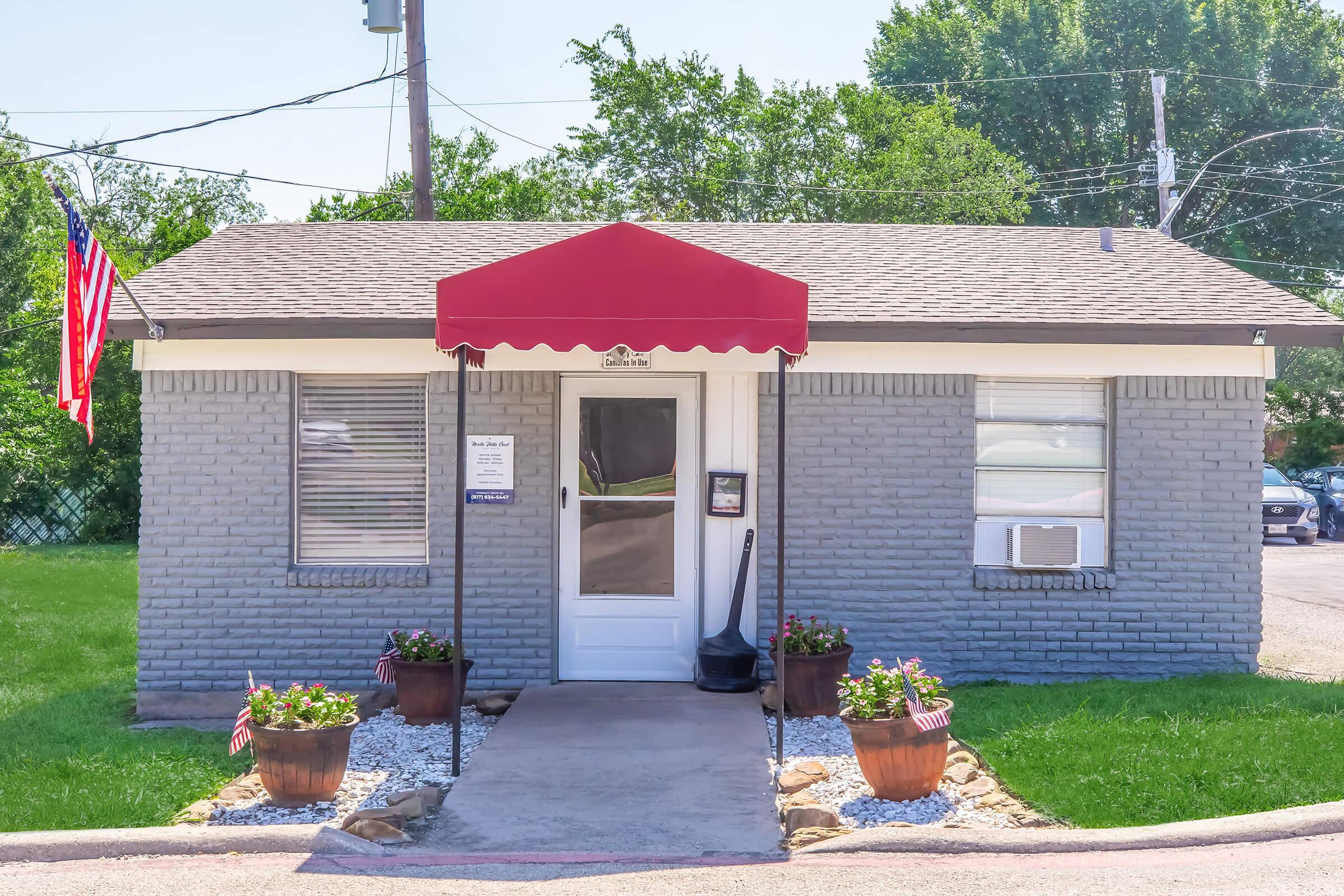 A small gray brick building with a red awning at the entrance. Two potted plants with flowers are on either side of the door, which is white. An American flag hangs beside the door, and there is an air conditioning unit on the wall. The surrounding area is grassy, and a parking lot is visible in the background.