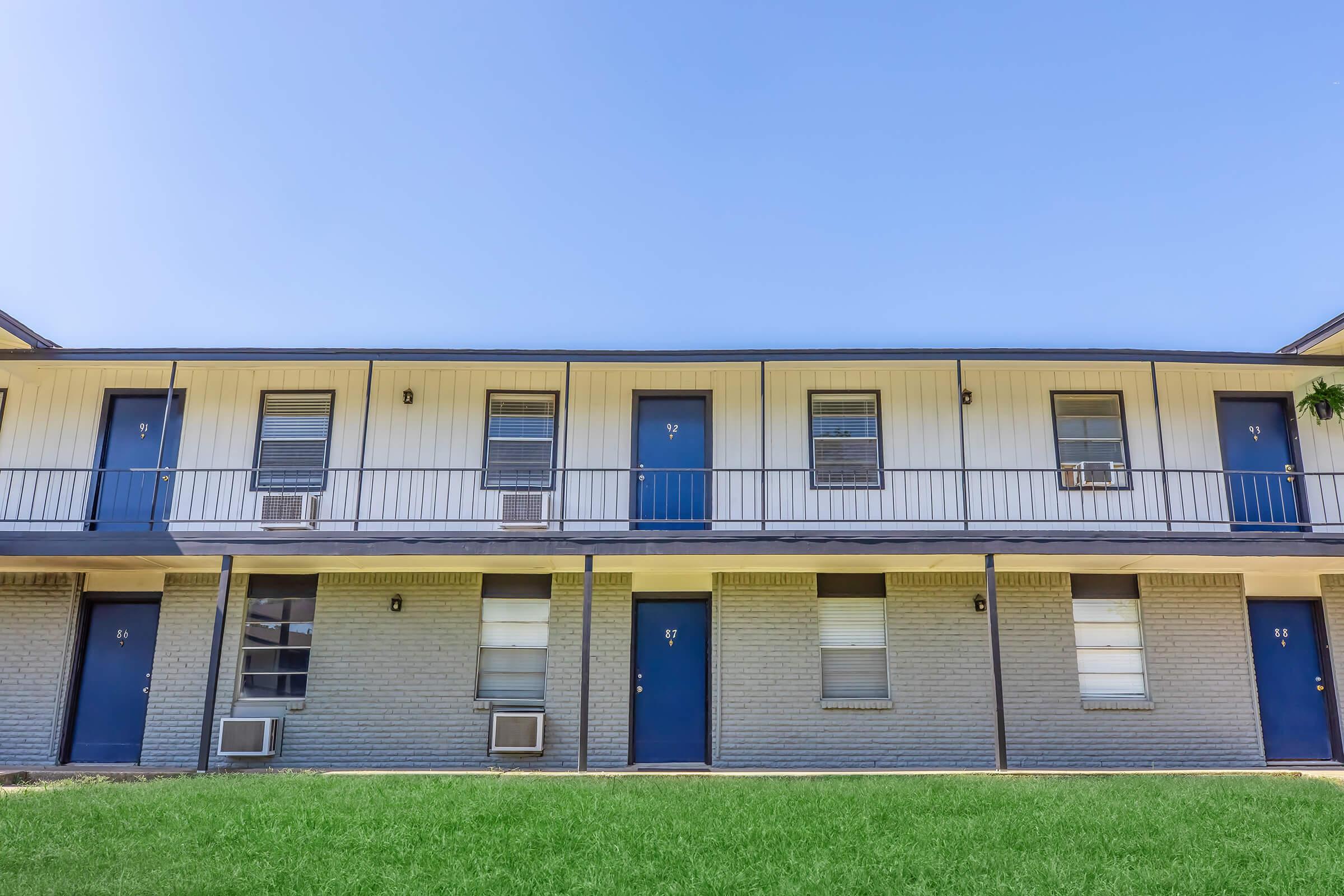 Two-story apartment building with multiple units, featuring blue doors and windows. The exterior is primarily gray brick with a white upper section. Each unit has an air conditioning unit installed beneath the windows. A grassy area is in the foreground under a clear blue sky.