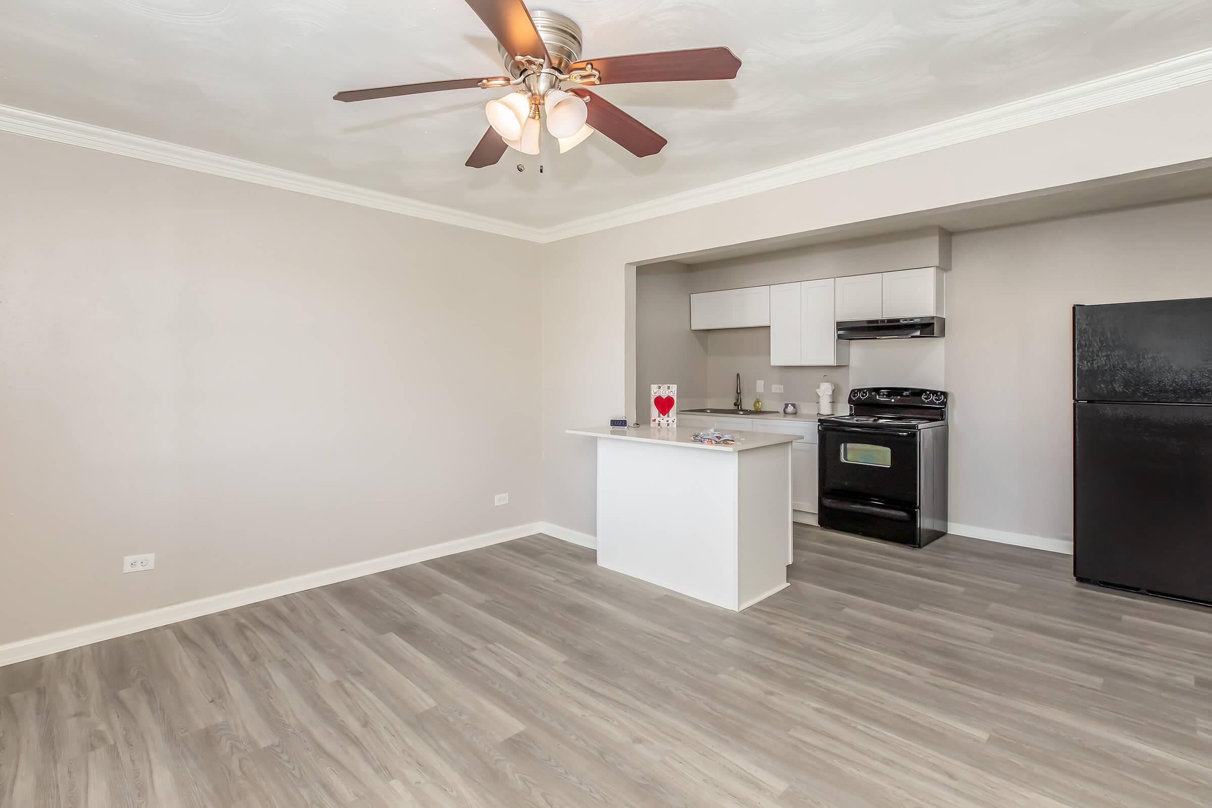 Interior view of a modern kitchen and living area featuring light-colored walls, wooden flooring, a ceiling fan with lights, and a black refrigerator. A small island separates the kitchen from the living space. The kitchen includes white cabinets and a stainless steel stove.