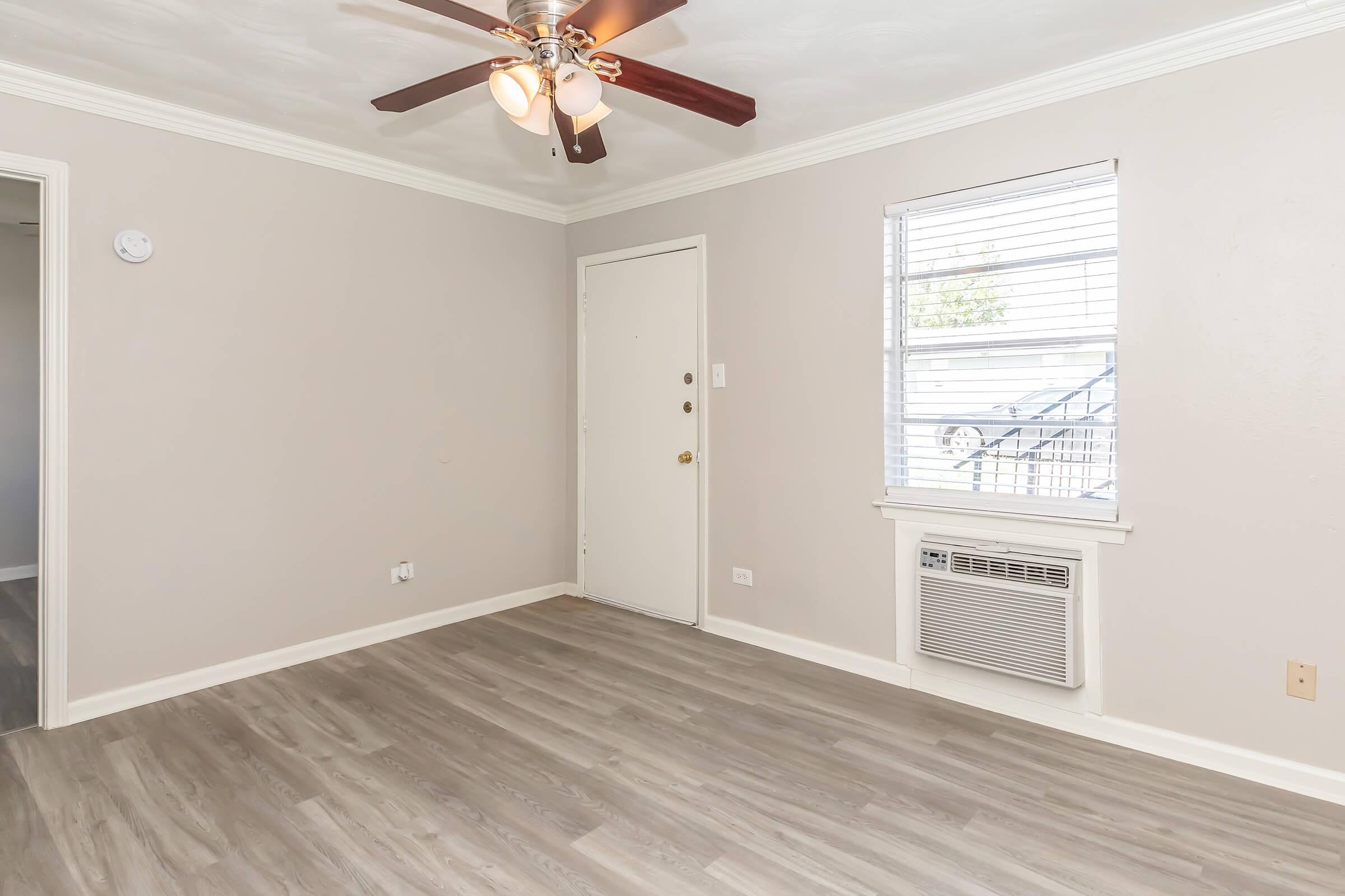 Spacious living room with light gray walls, a ceiling fan, and a window with white blinds. There is a white door leading outside, an air conditioning unit, and a light-colored laminate floor. The room is well-lit, creating a welcoming atmosphere.