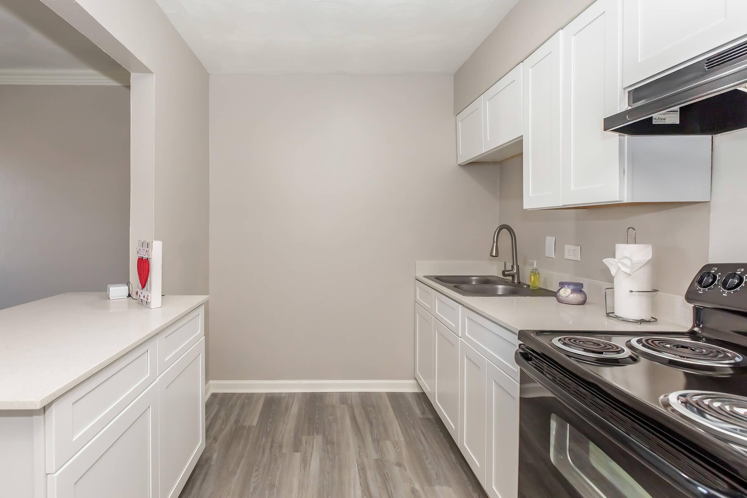 A modern kitchen featuring white cabinetry, a countertop, a black stove, and stainless steel appliances. The walls are painted in a light beige color, and the flooring is a gray wood-like material. A small decorative item is visible on the countertop, adding a touch of warmth to the space.