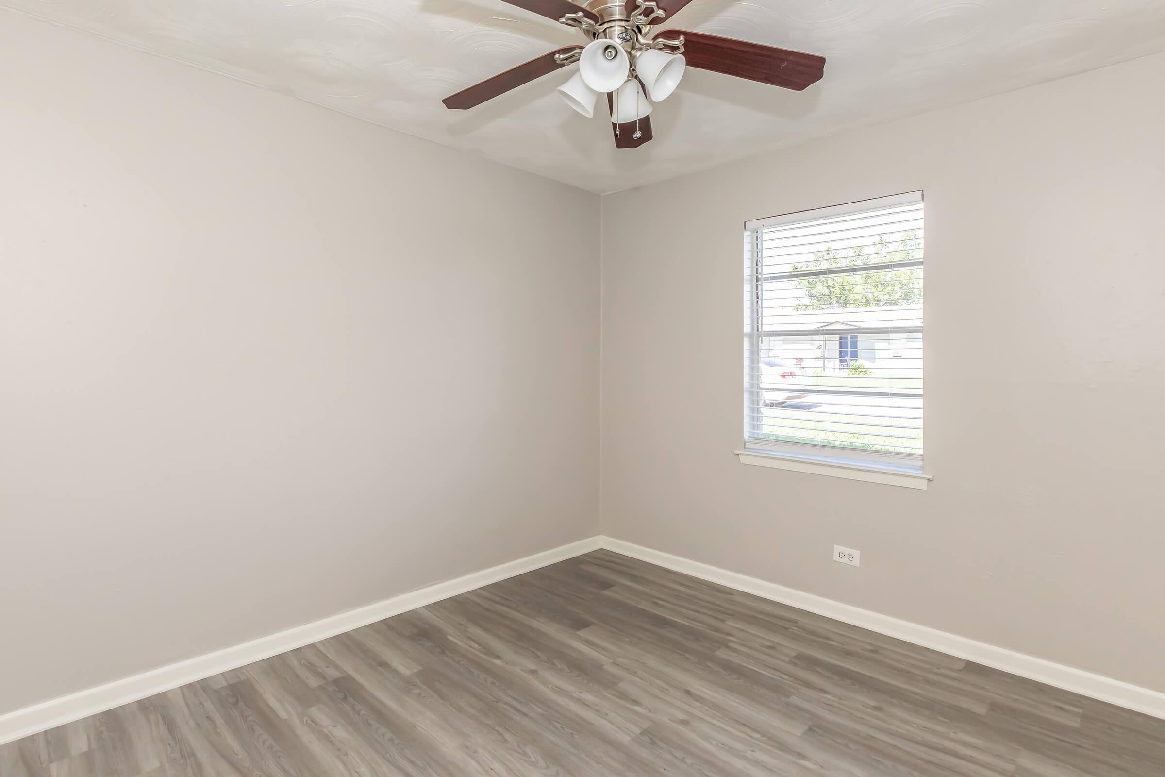 A well-lit, empty room with light gray walls and a ceiling fan featuring five blades. The floor is covered in wood-like laminate. There is a window with blinds, allowing natural light to enter and offer a view of the exterior. The overall atmosphere is clean and spacious, suitable for various uses.