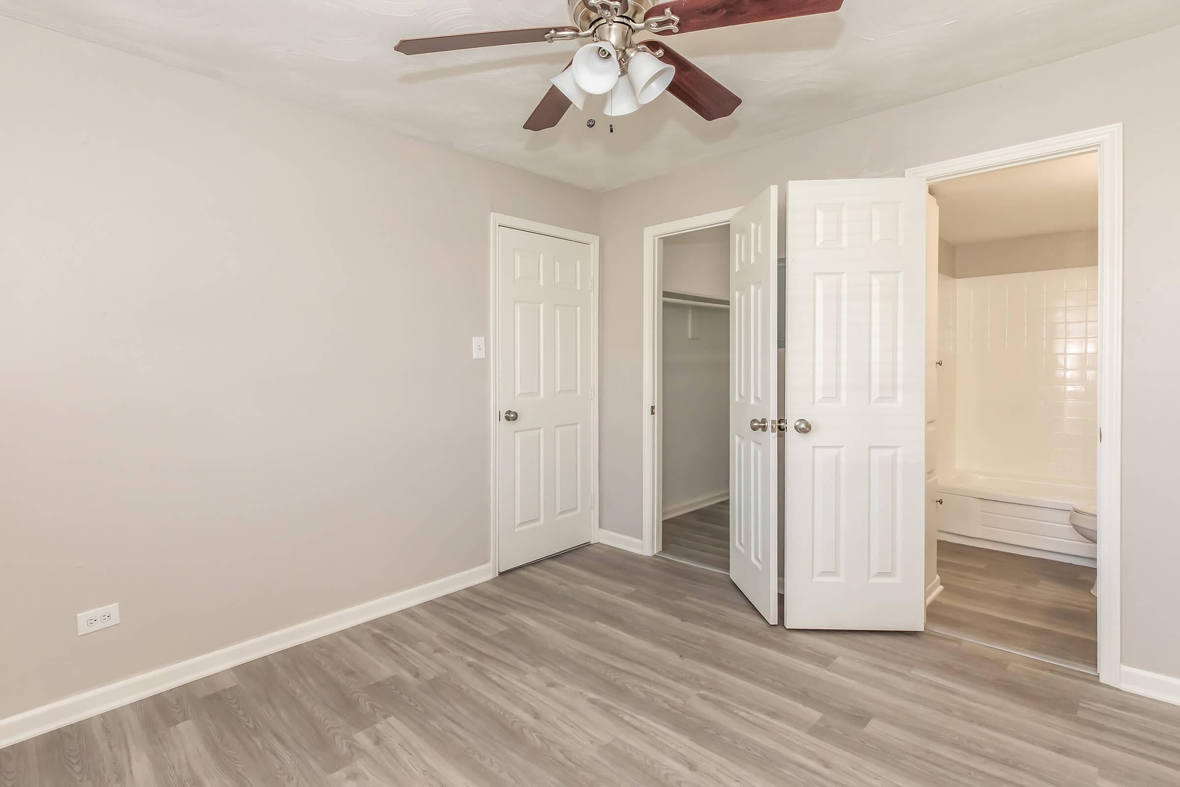 A well-lit, empty room featuring light-colored walls and new flooring. Two closed doors lead to a closet and a bathroom. A ceiling fan with wooden blades is visible, adding to the room's modern appeal. The overall atmosphere is spacious and inviting, ready for personal touches.