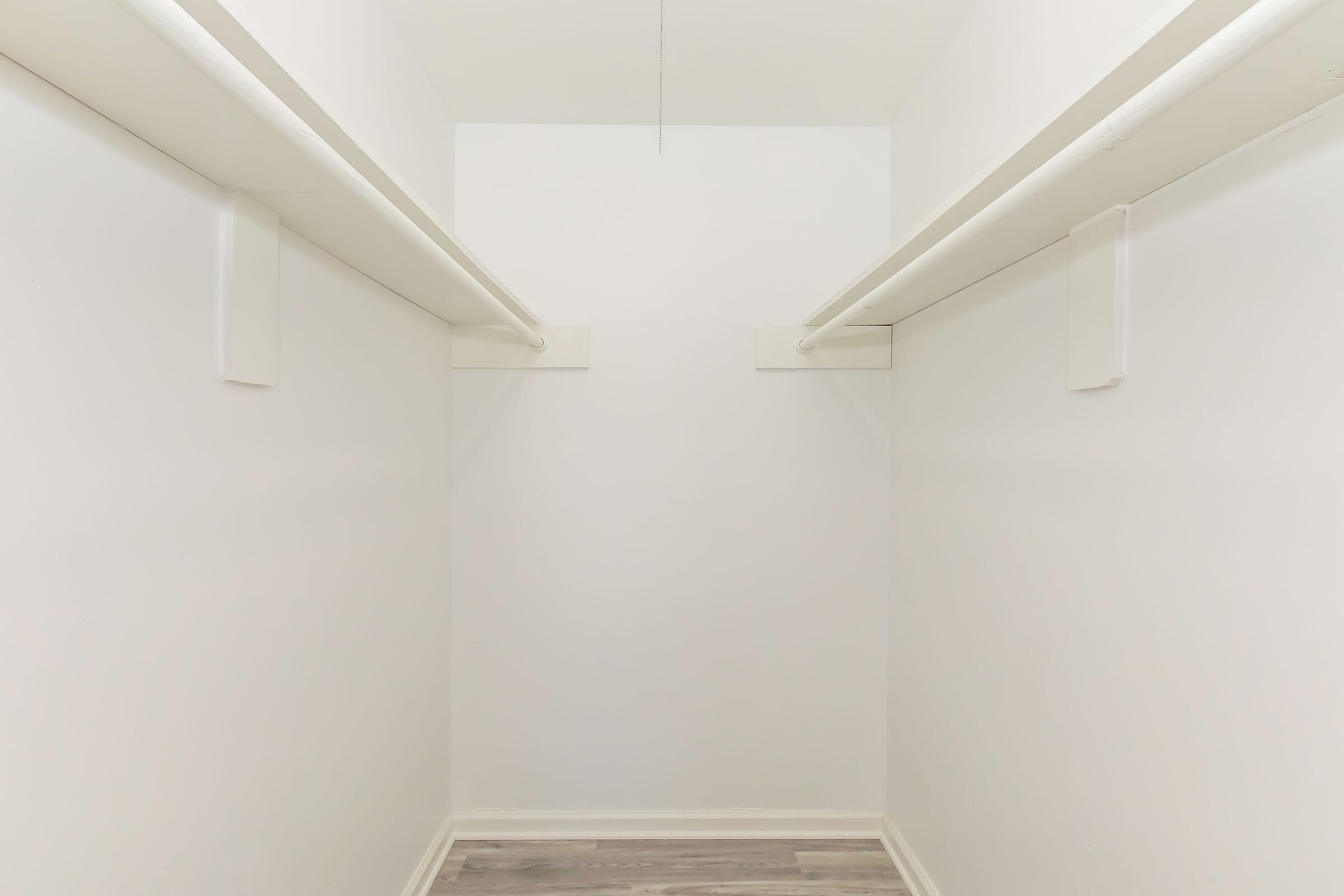 A spacious empty closet with white walls and two horizontal rods for hanging clothes. The floor is light-colored, and there is no visible storage or items present.