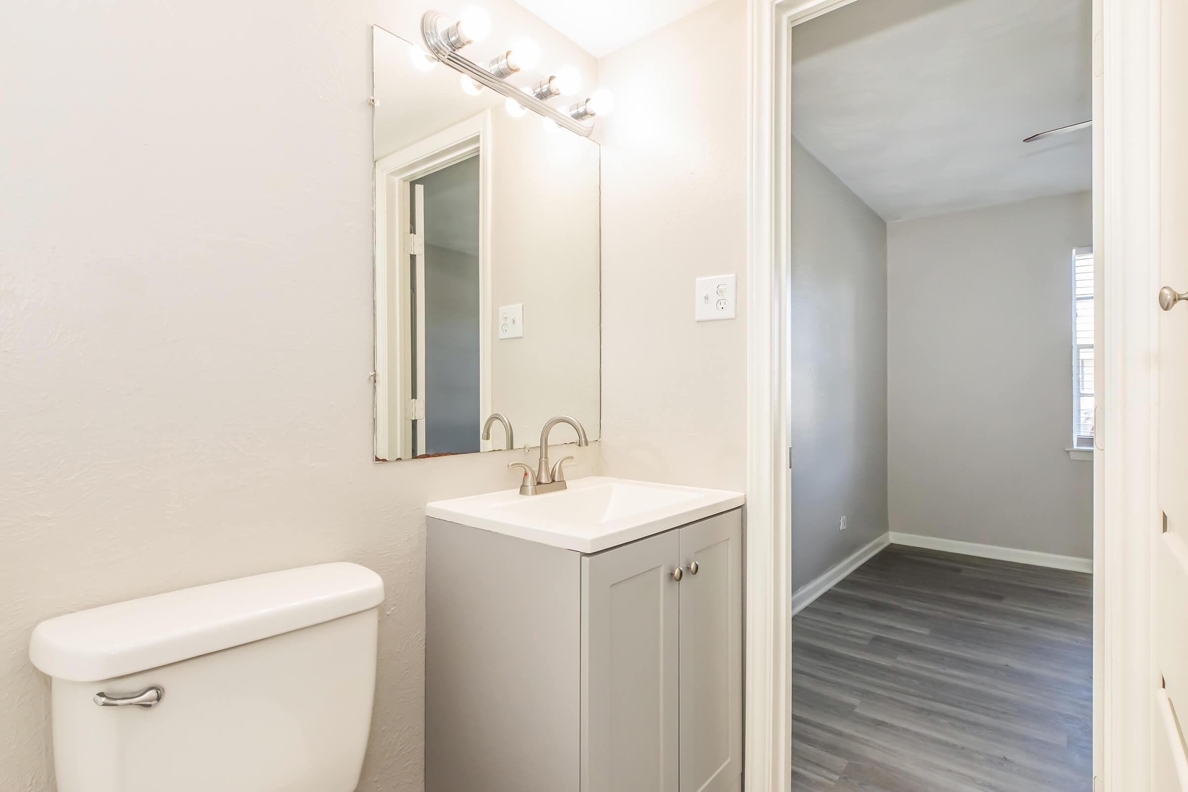 A small bathroom featuring a modern sink with a mirror above it, and a toilet nearby. The walls are painted light neutral tones, and the flooring is a light wood laminate. In the background, a doorway leads to a brightly lit room with a window.