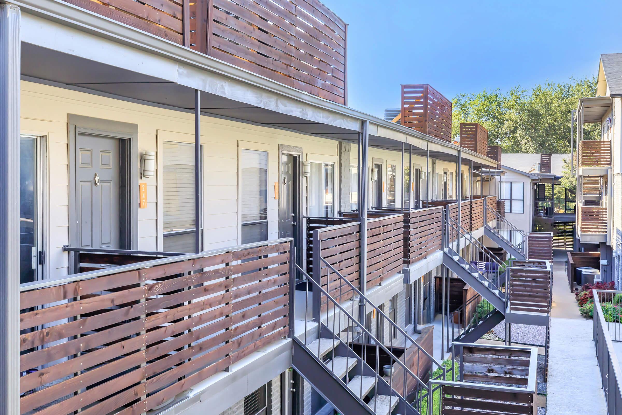 A view of a modern apartment complex featuring multiple units with wooden balconies and metal stairs. The architecture includes light-colored walls and a clean, contemporary design. Lush greenery is visible in the background, contributing to a pleasant and inviting atmosphere.