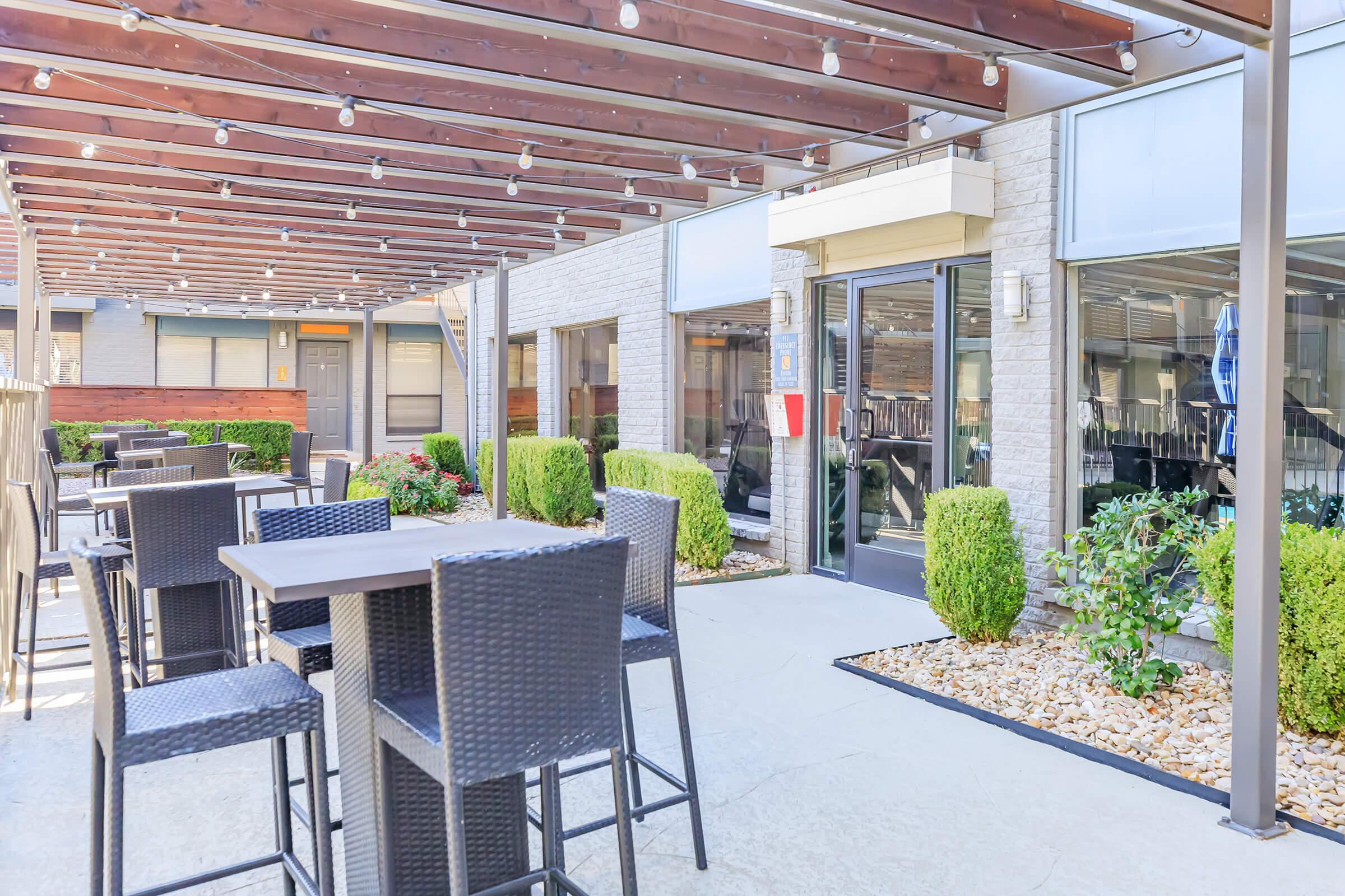An outdoor dining area with modern furniture, featuring dark woven tables and chairs under a wooden pergola. The space is well-lit with string lights and has neatly arranged shrubs and gravel landscaping. Large glass doors lead into a building, enhancing the inviting atmosphere.