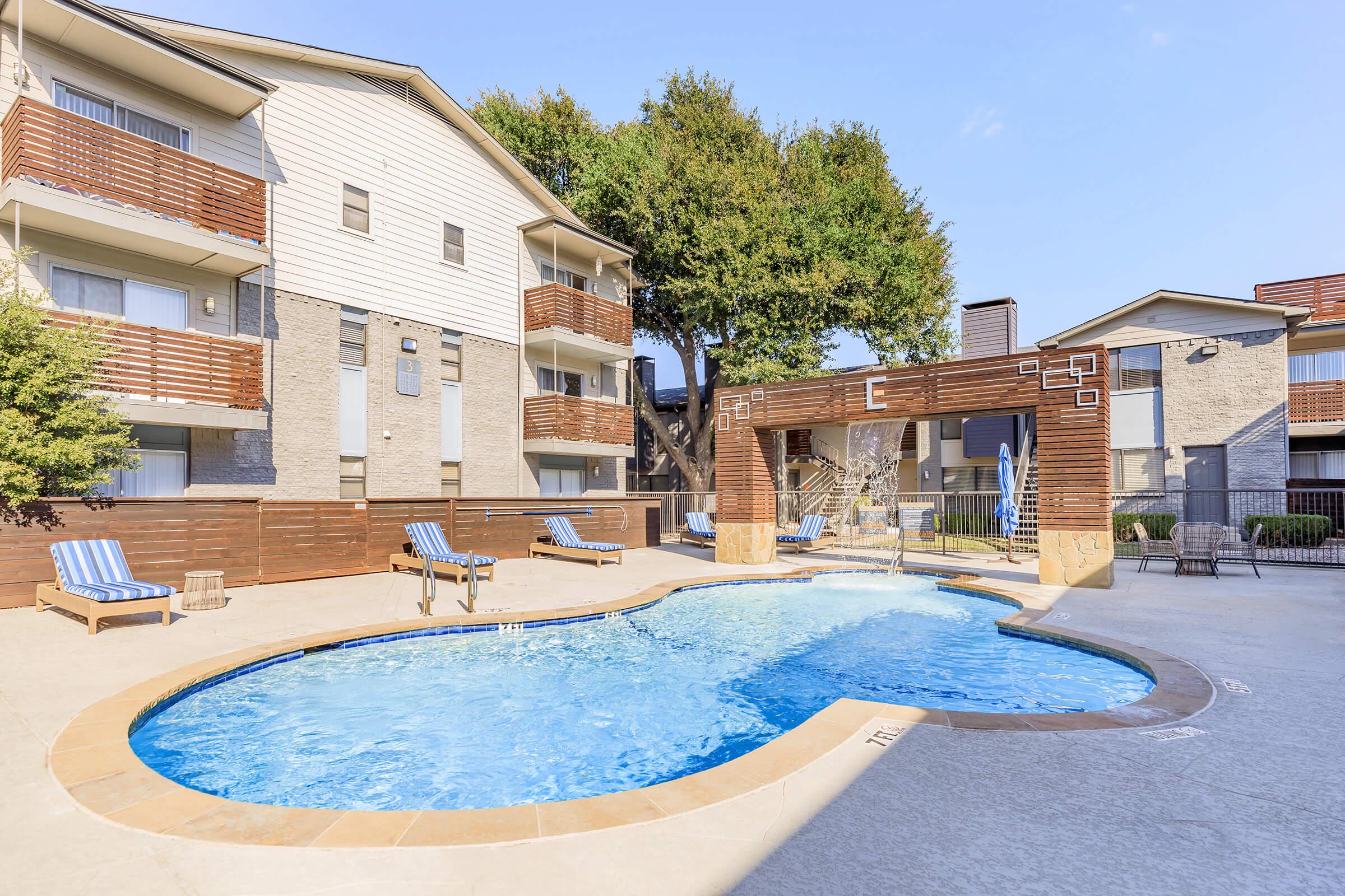 A clear blue swimming pool surrounded by lounge chairs in a well-maintained apartment complex. The area is landscaped with greenery, and the buildings feature a modern design with balconies. The sky is bright and sunny, creating a welcoming outdoor atmosphere.