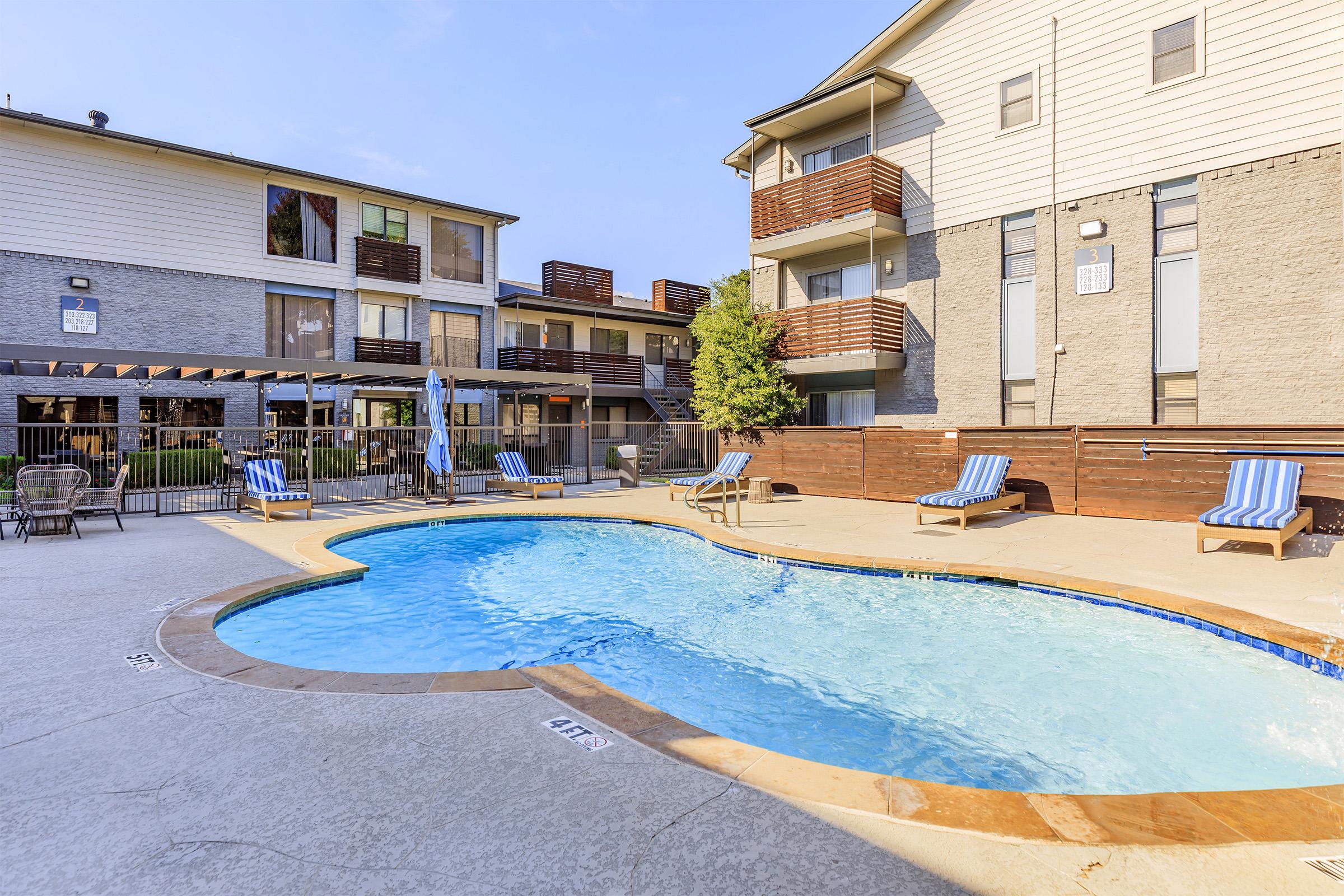 A swimming pool surrounded by lounge chairs and umbrellas, set in a residential complex with multi-story buildings in the background. The pool features a curved shape and is situated on a sunlit patio area, perfect for relaxation and leisure activities.