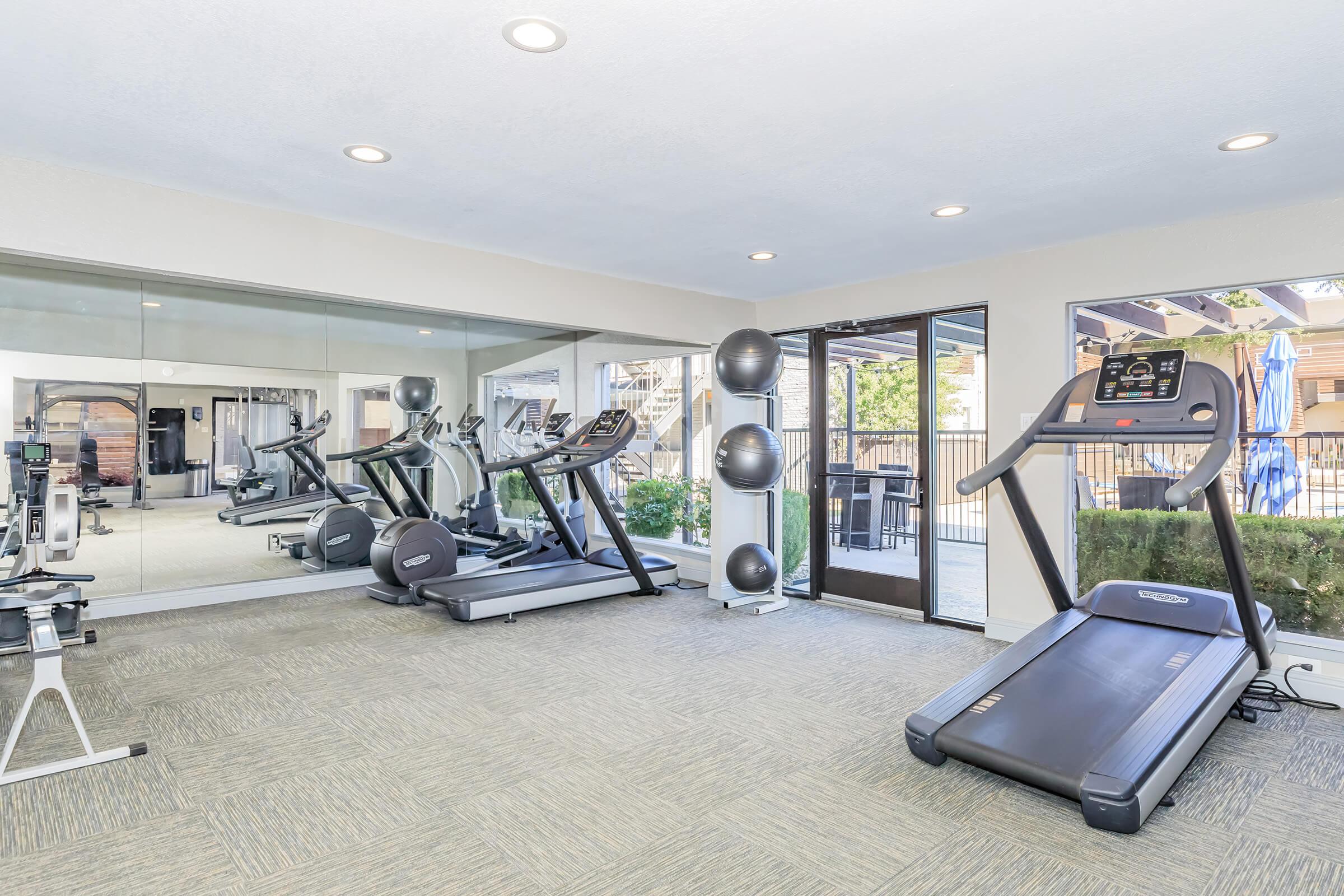 A modern gym interior featuring multiple exercise machines, including treadmills and stationary bikes. The space is well-lit and includes a large mirror on one wall. Large windows offer views of an outdoor area with greenery and poolside amenities.