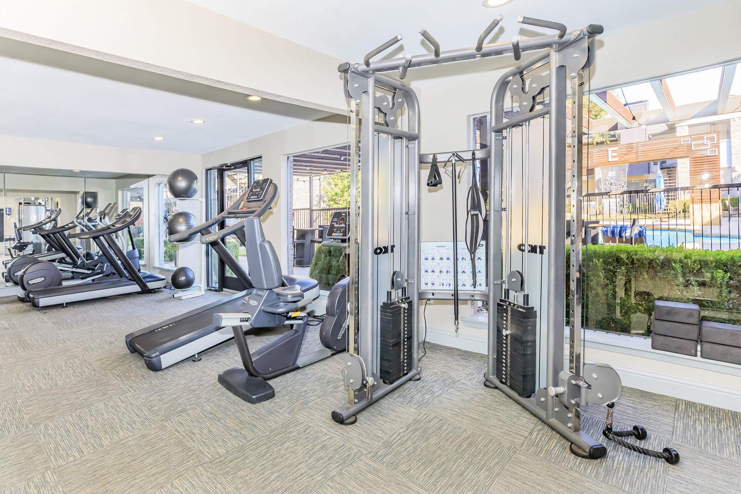 A modern gym interior featuring various exercise equipment, including a cable machine, treadmill, and elliptical. Brightly lit with large windows, the space also includes fitness balls and a wall chart. The setting is clean and inviting, ideal for workouts and fitness routines.