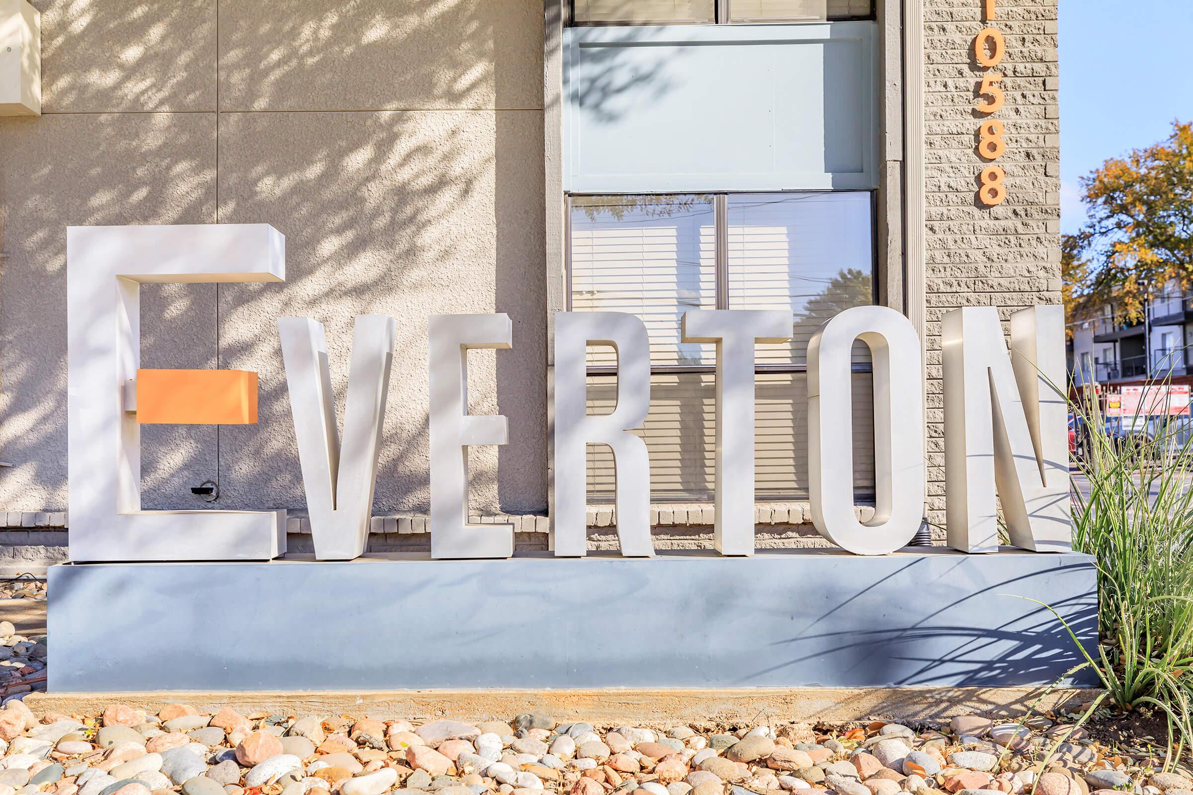 A large, stylized sign displaying the word "Everton." The letters are primarily white, with a single orange accent. The sign is positioned in front of a building, surrounded by small stones and grass, with sunlight casting shadows on the wall behind it.