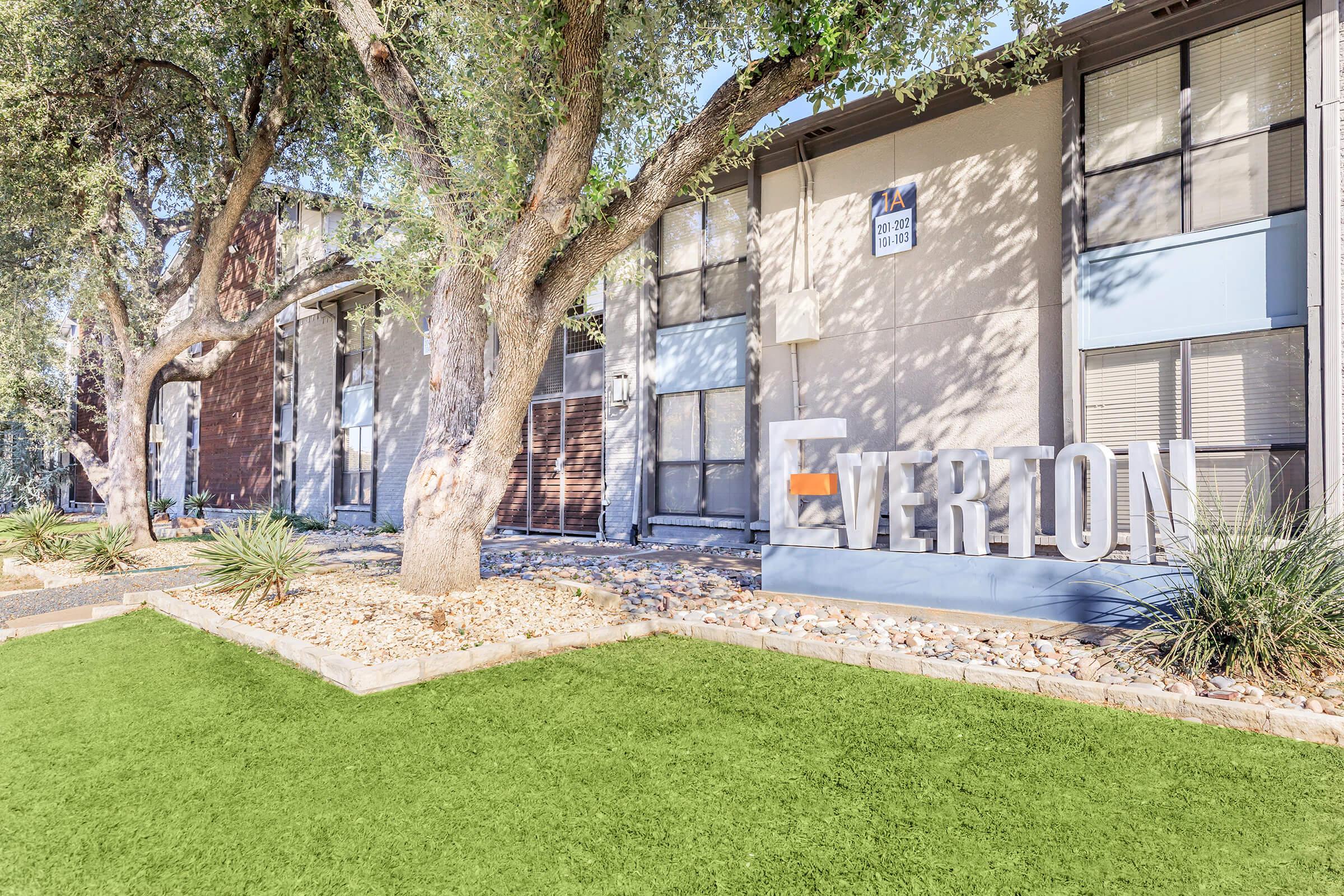 A landscaped area featuring a large sign that reads "EVERTON," surrounded by trees and shrubs. The background shows a modern building with multiple windows and a manicured lawn. The scene conveys a welcoming atmosphere, suitable for an apartment complex or residential community.